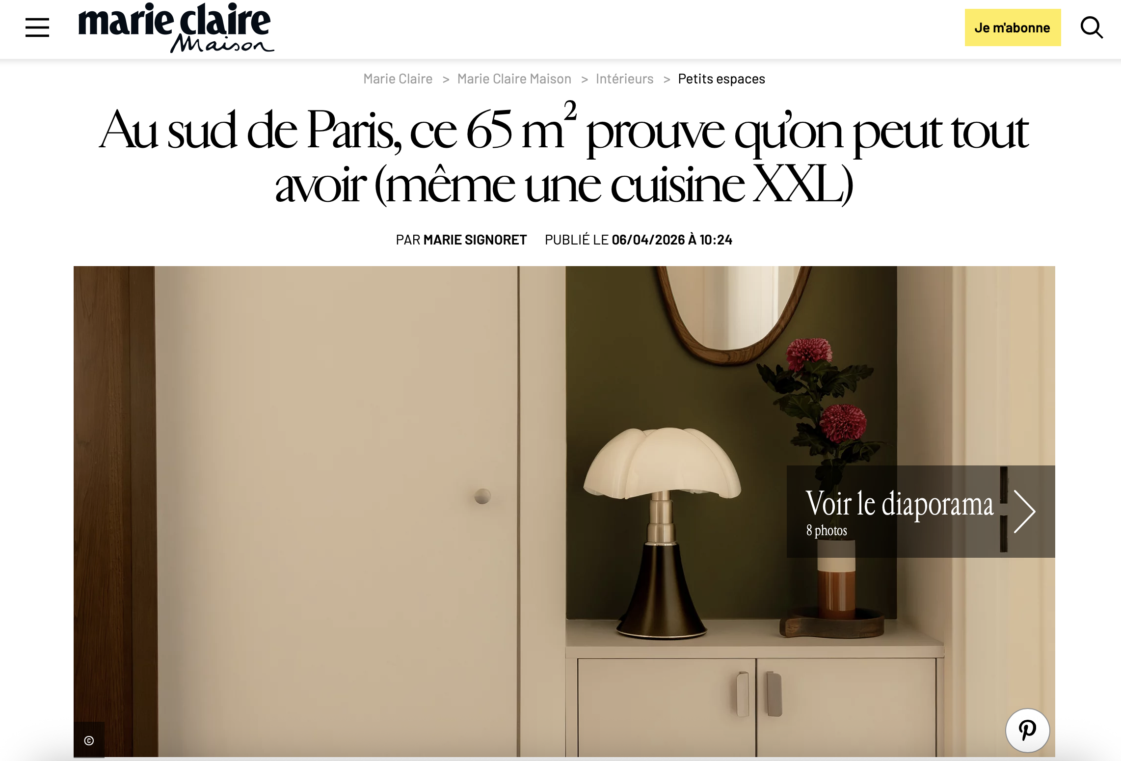 Une lampe de table sur une commode blanche, un miroir rond au mur, une plante florale avec des fleurs roses, un vase dans un plateau en bois, et une partie d'un mur beige et vert.