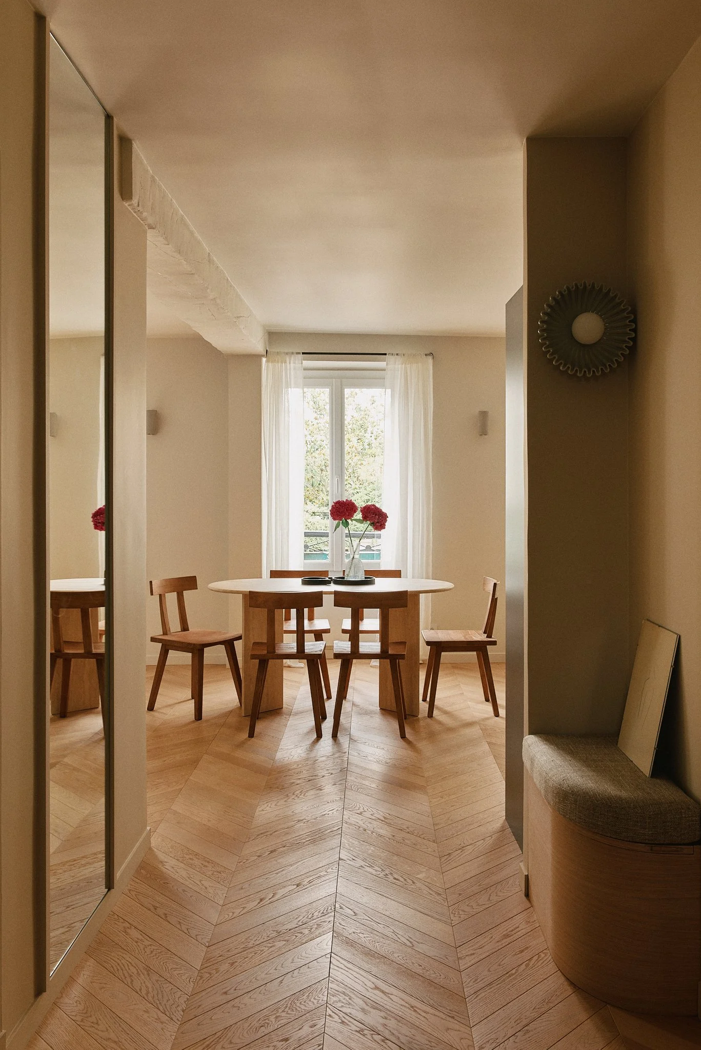 Salle à manger avec table ronde en bois, six chaises en bois, une coupelle avec des fleurs roses, fenêtre avec rideaux blancs, sol en parquet en chevrons.