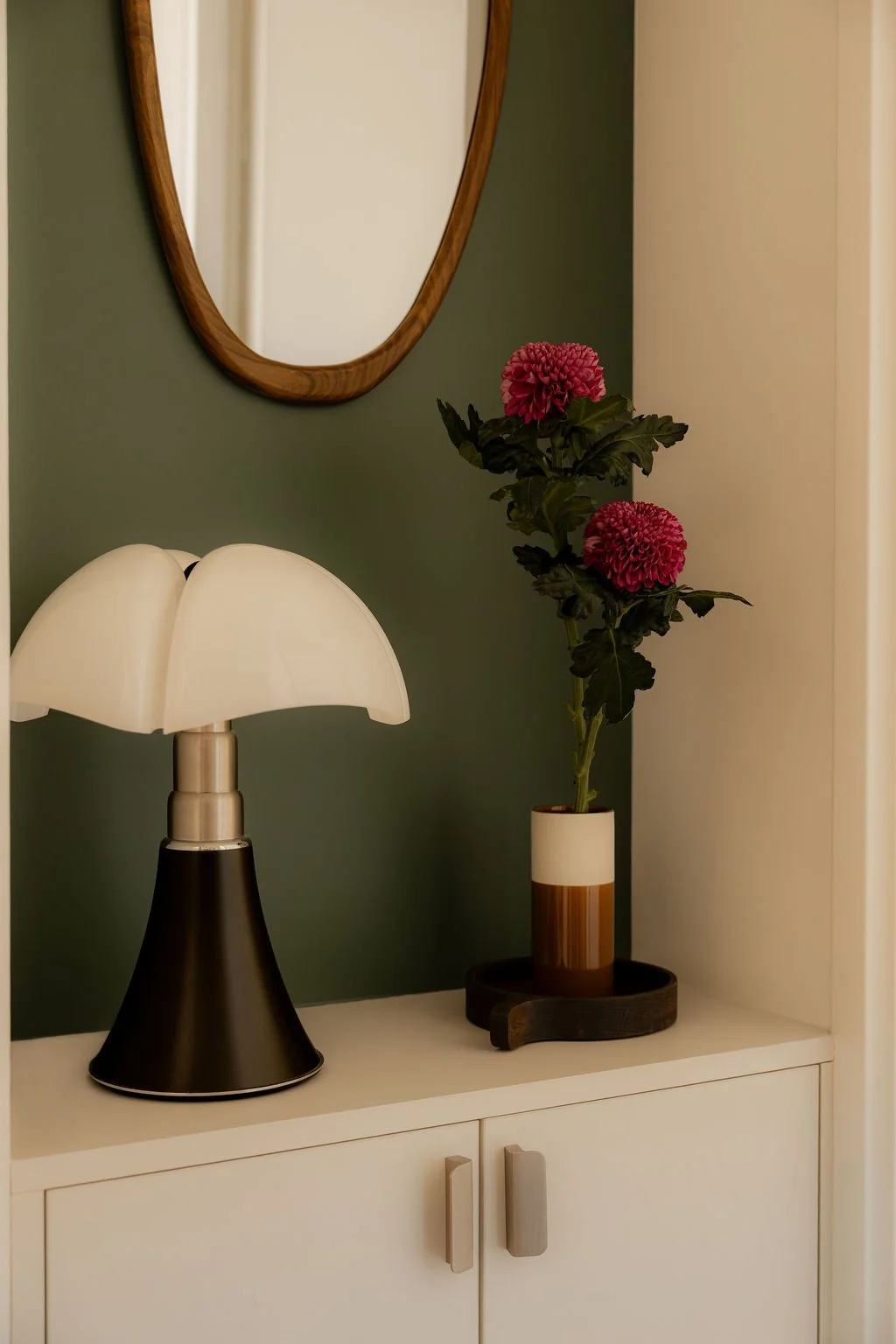 Une lampe design, un miroir en bois et une plante avec des fleurs roses dans un vase sur une commode