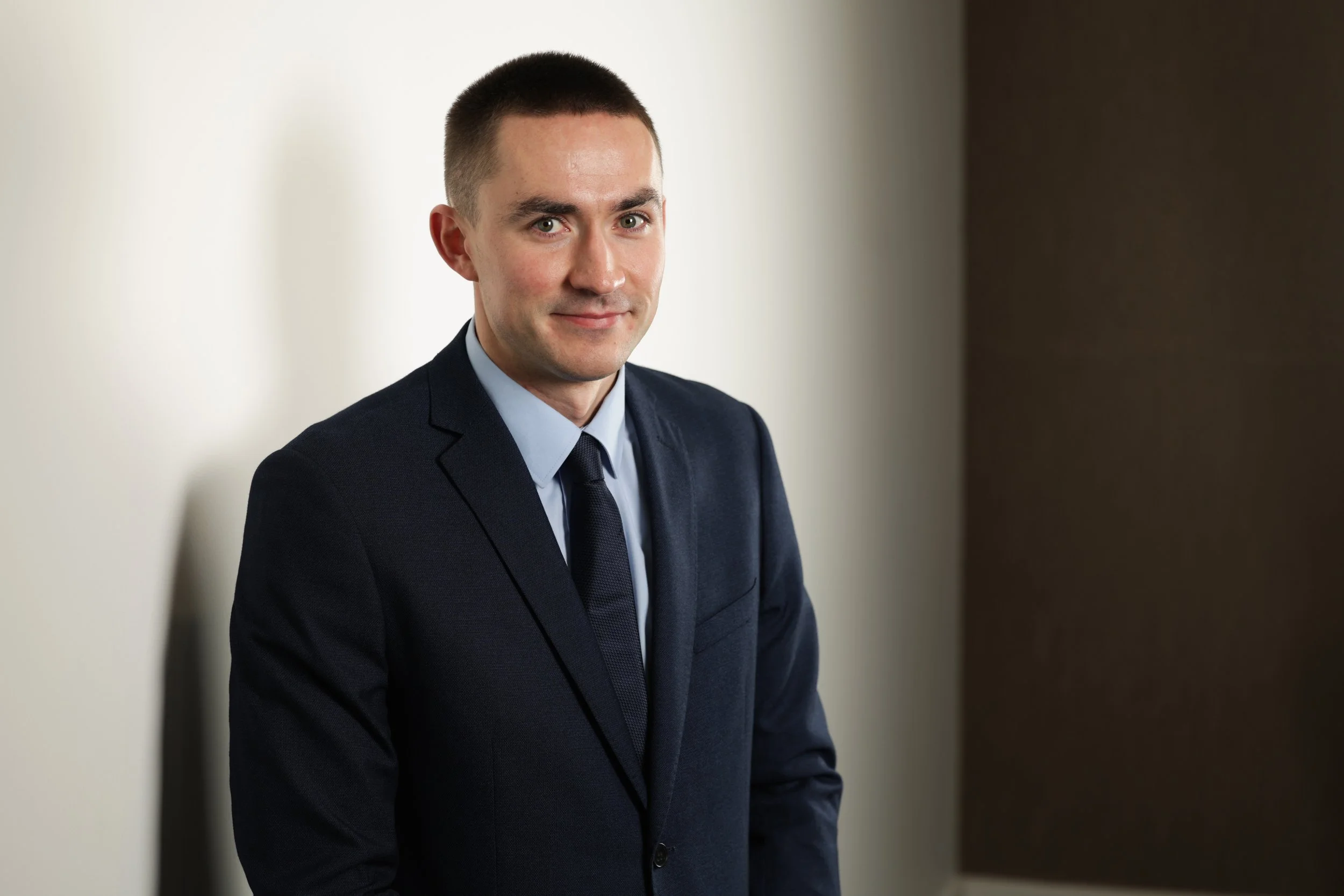 Portrait of a young man with short dark hair and light skin, wearing a dark suit, light blue shirt, and dark tie, smiling softly indoors against a simple background.