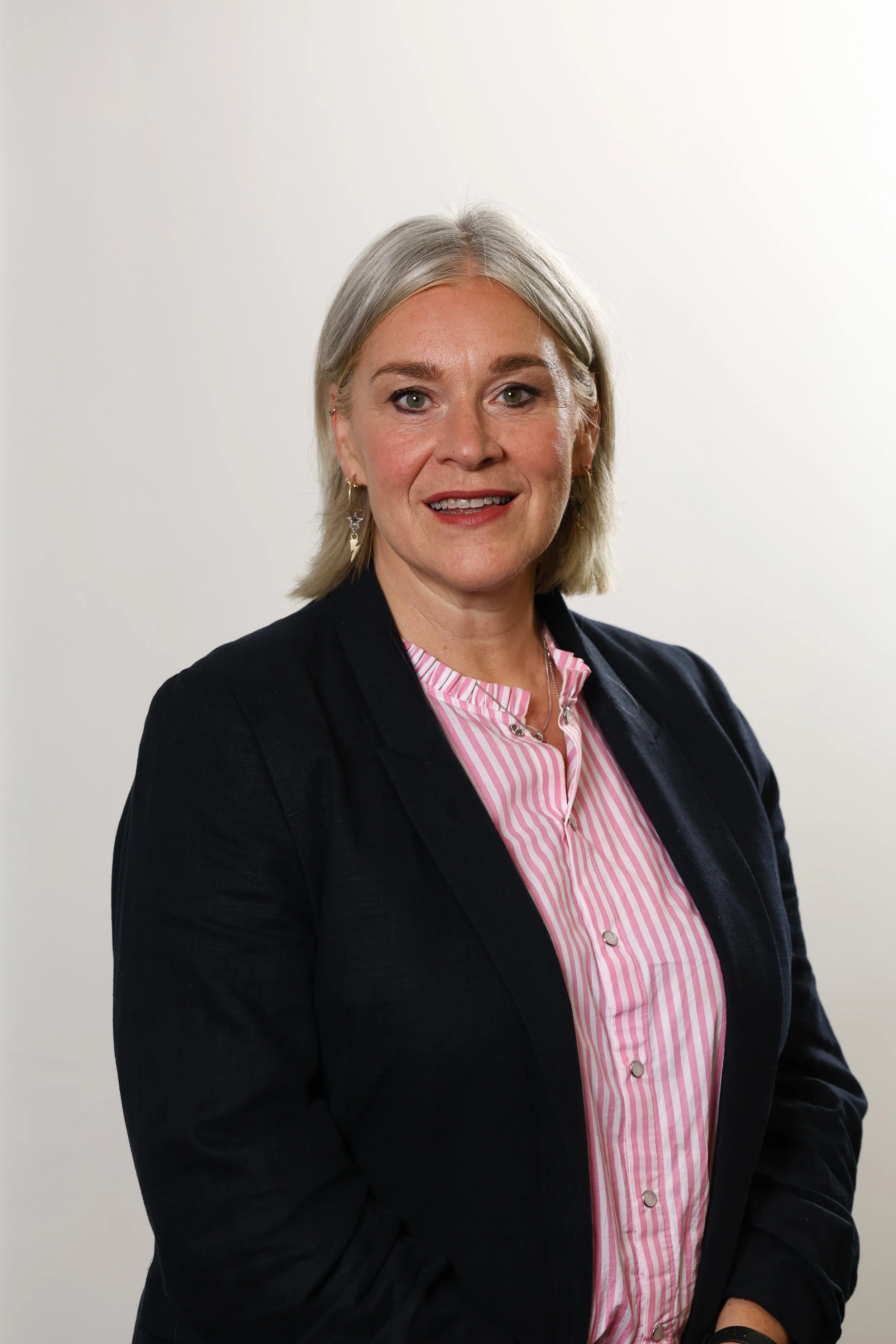 A woman with gray hair, wearing a black blazer over a pink and white striped shirt, smiling and looking at the camera against a plain white background.