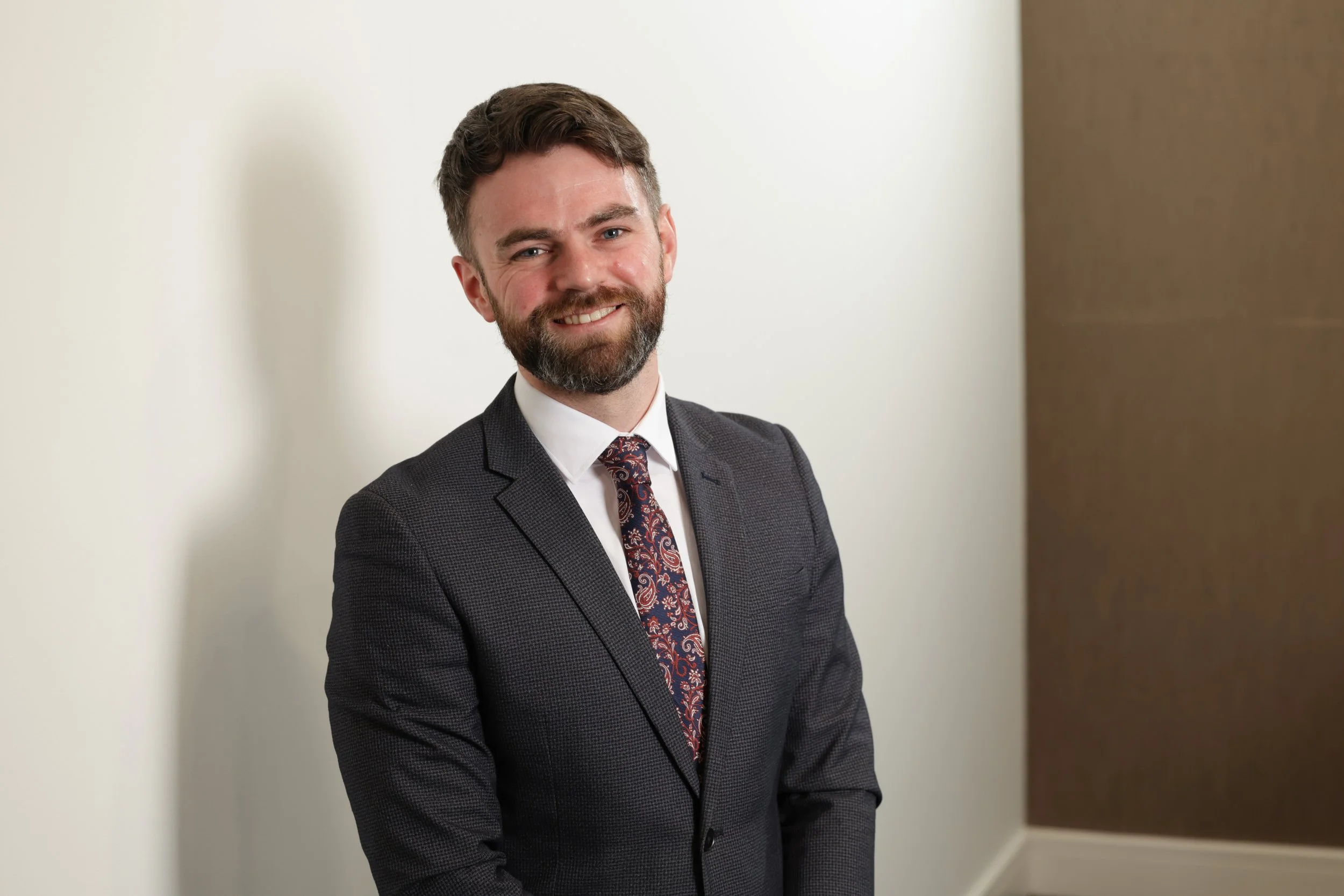 A man with a beard and brown hair, wearing a dark grey suit and a paisley tie, smiling and standing against a white wall with a brown panel.