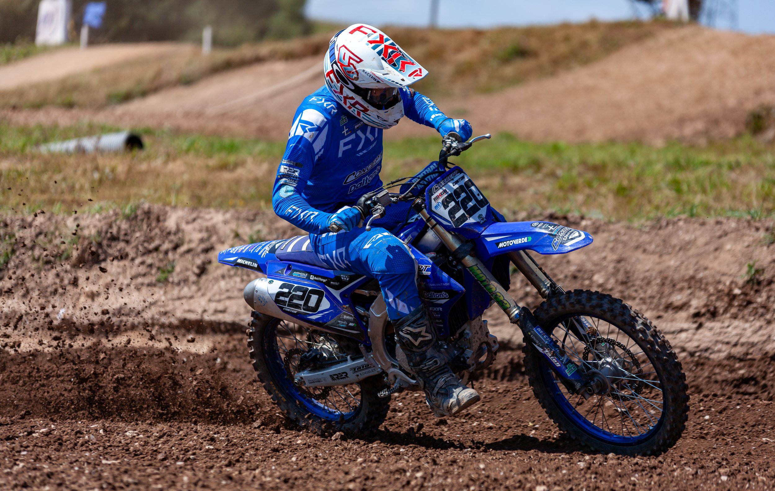 A motocross rider in a blue racing suit and helmet riding a blue dirt bike on a dirt track, producing a spray of dirt as the bike leans into a turn.