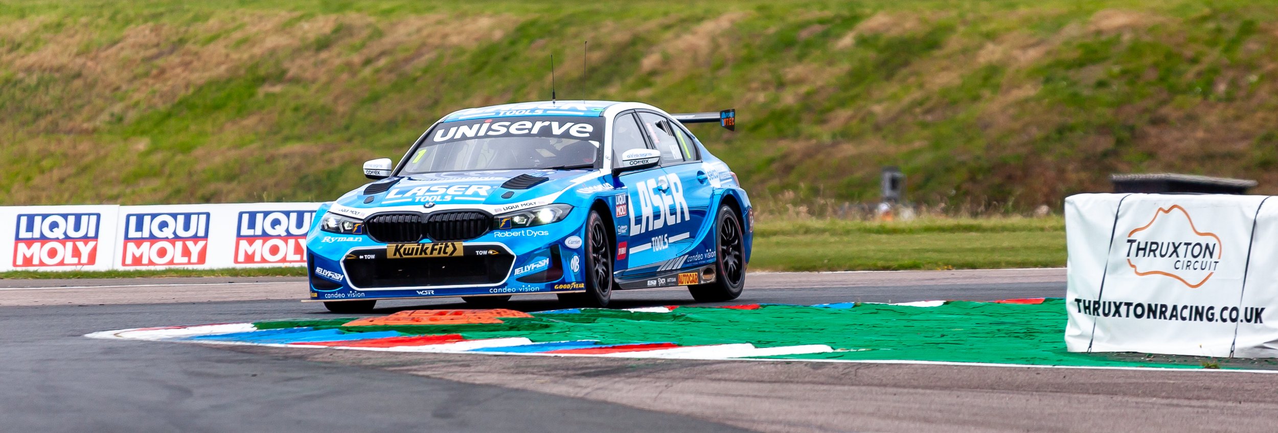 A blue race car on a track near a curb and advertising banners, at Thruxton Circuit.