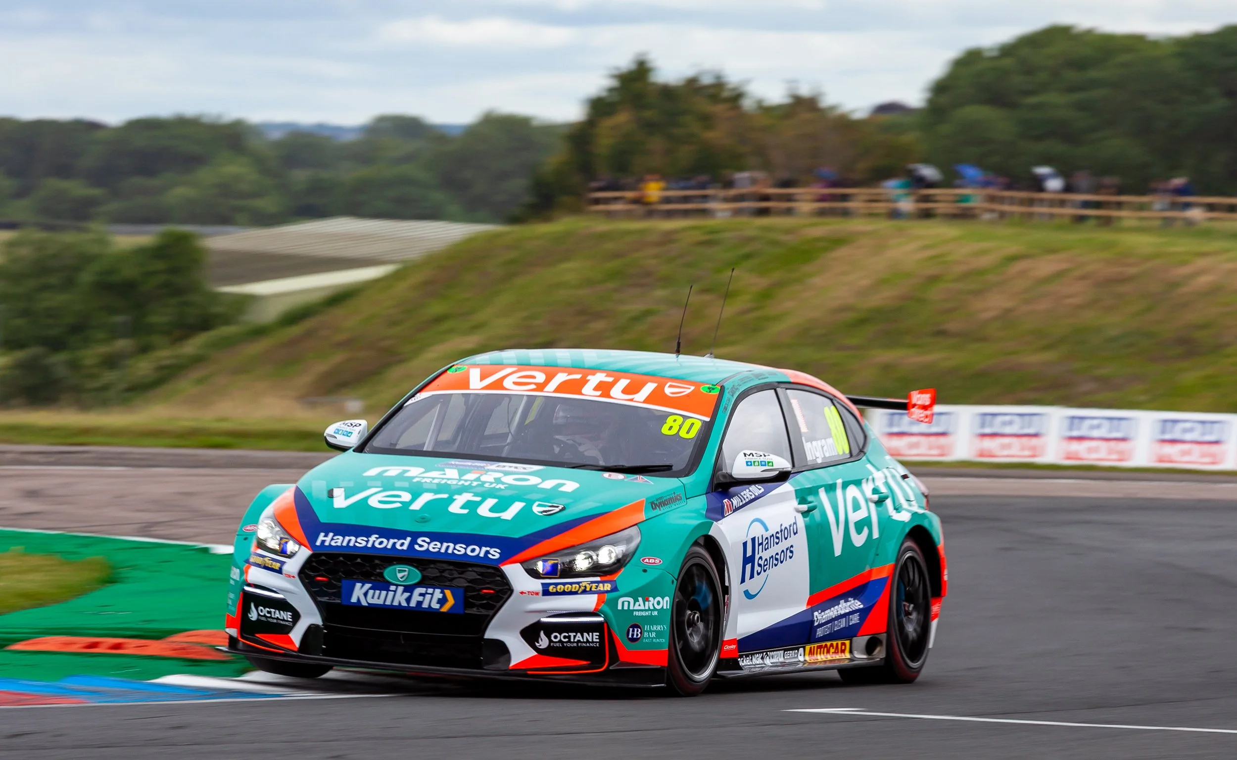 A race car with the logos Veru and Hansford Sensors rounding a bend on a racetrack with spectators in the background.