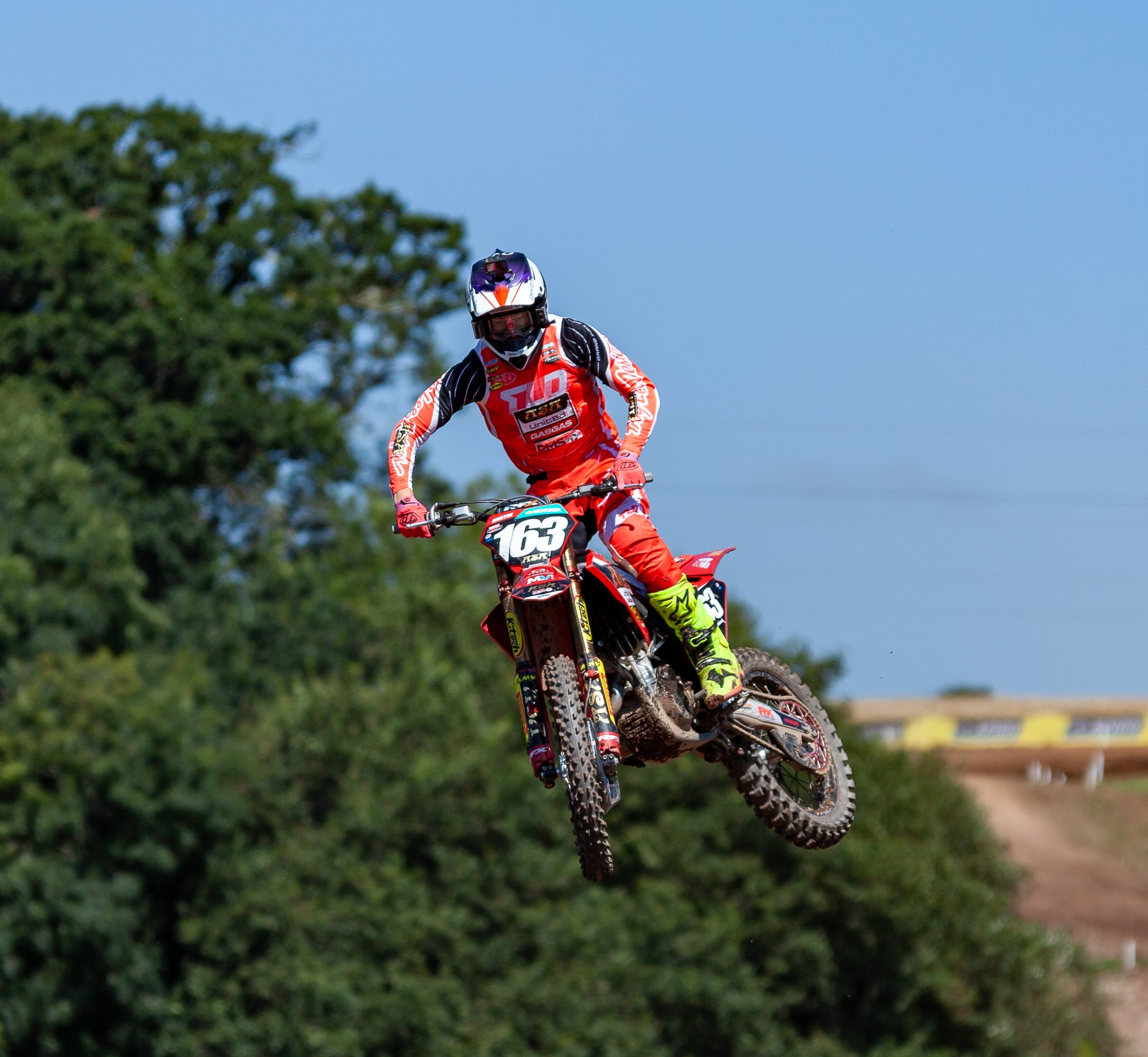 Motorcycle rider in red gear mid-air on dirt track with trees in background and cloudy sky.