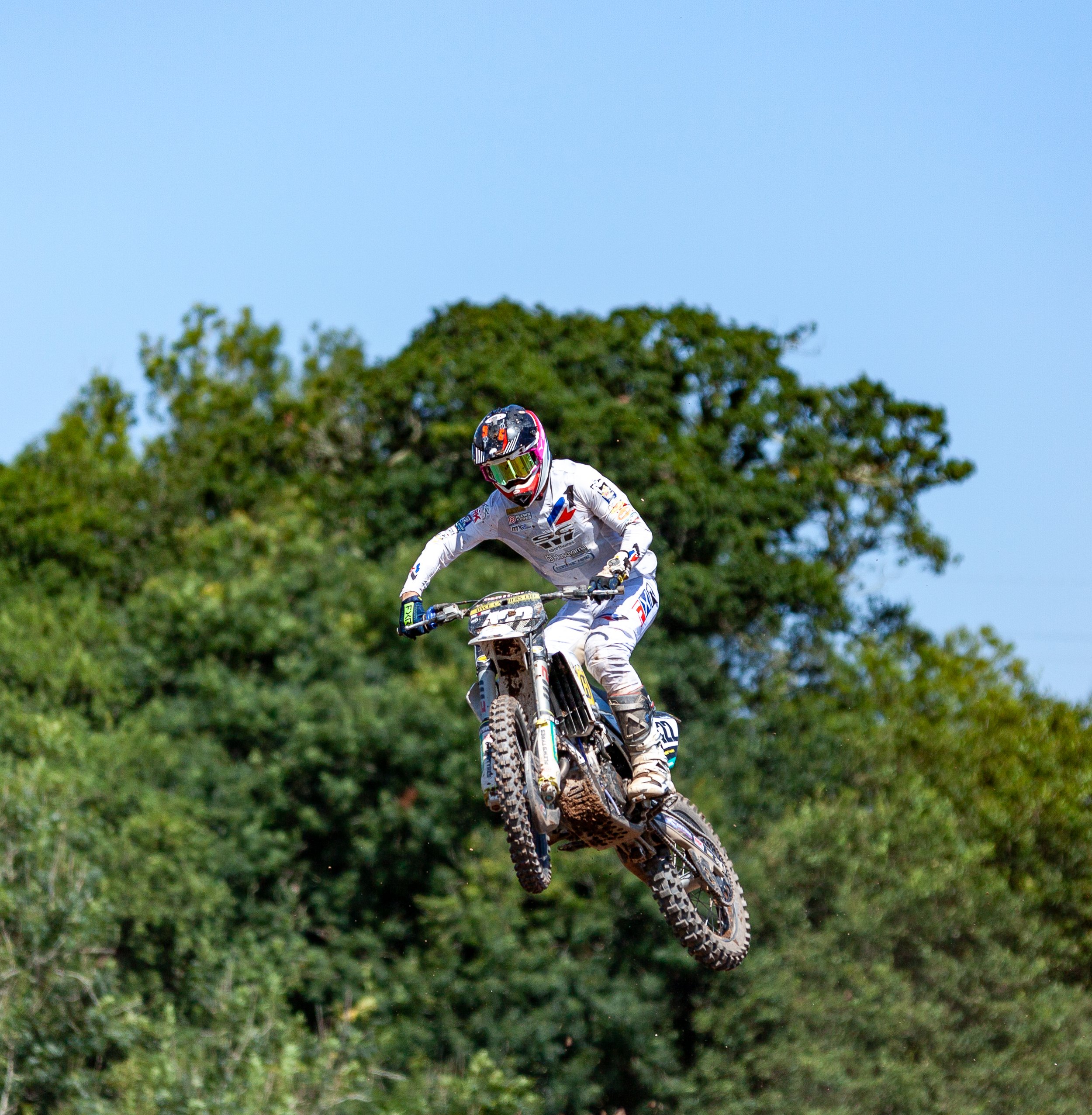 Motocross rider in a white suit and helmet mid-air on a dirt bike, with a green leafy background and blue sky.