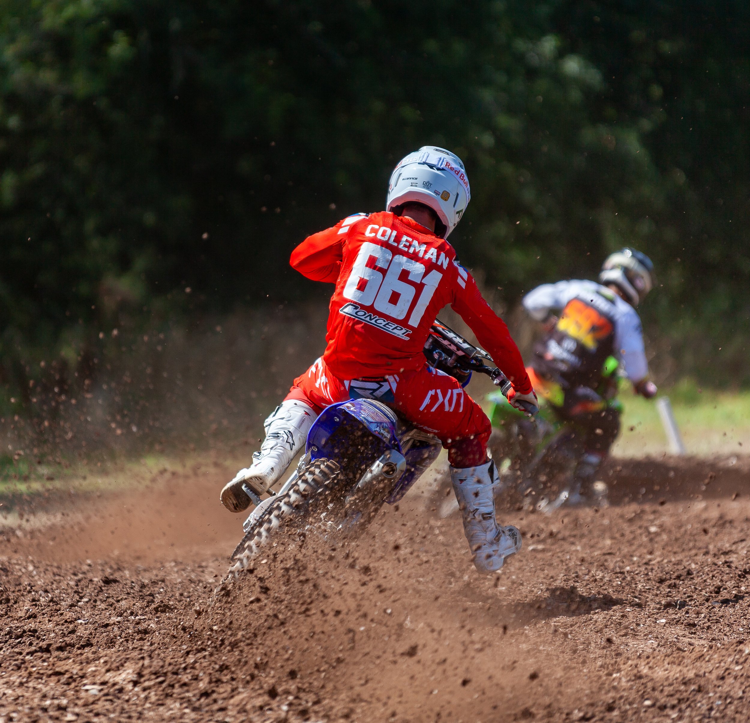 Two motocross riders in gear riding on a dirt track, kicking up dust, with a forested background.