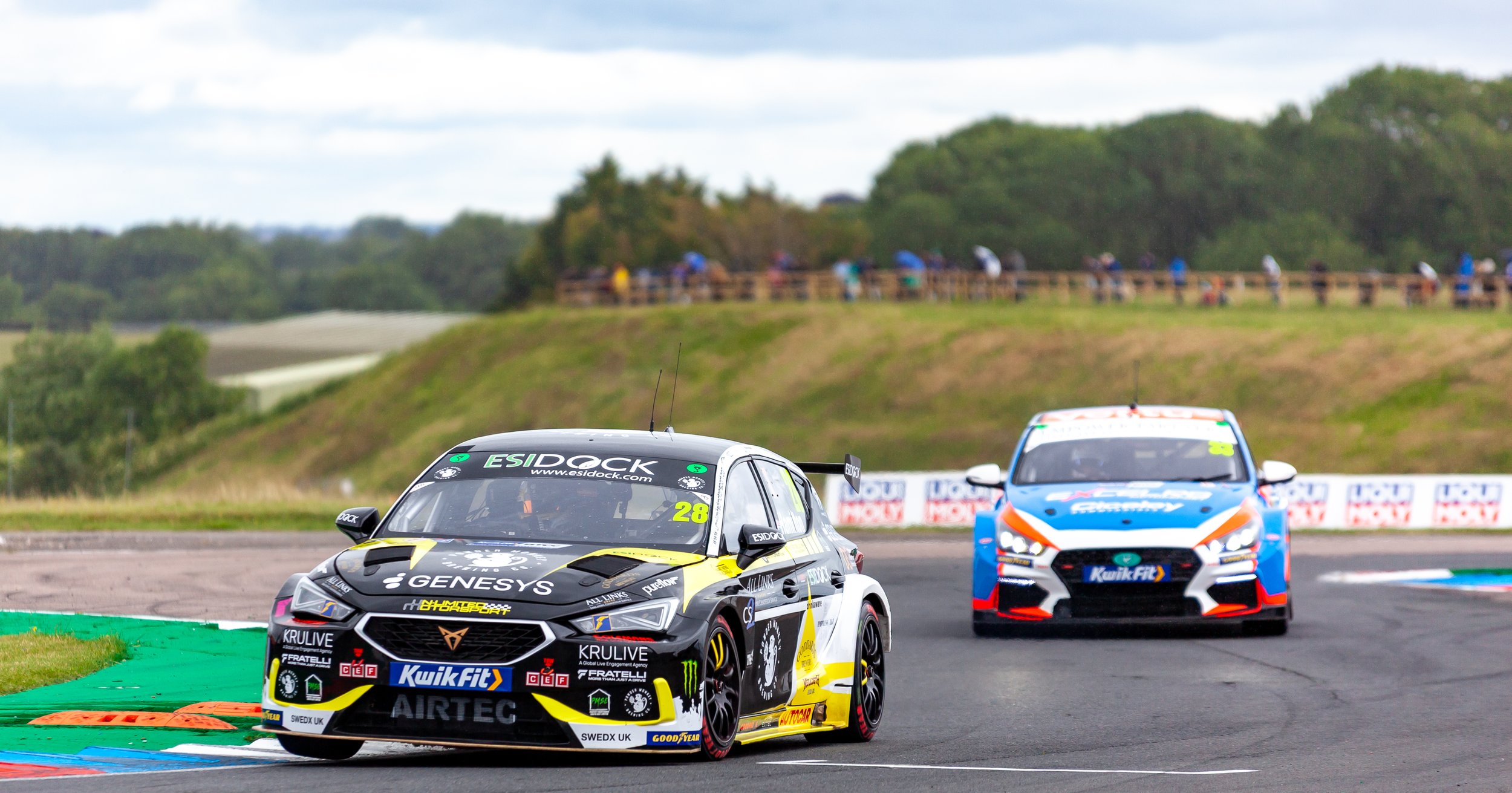 Two race cars on a racetrack, with the black and yellow car leading and the blue, white, and orange car trailing behind. Spectators are visible on a hill in the background.