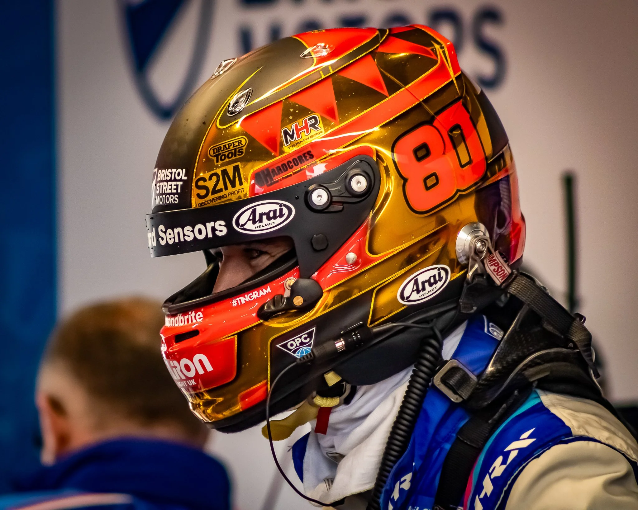 Race car driver wearing a colorful helmet with the number 80, racing gear, and earphones, in a racing environment.