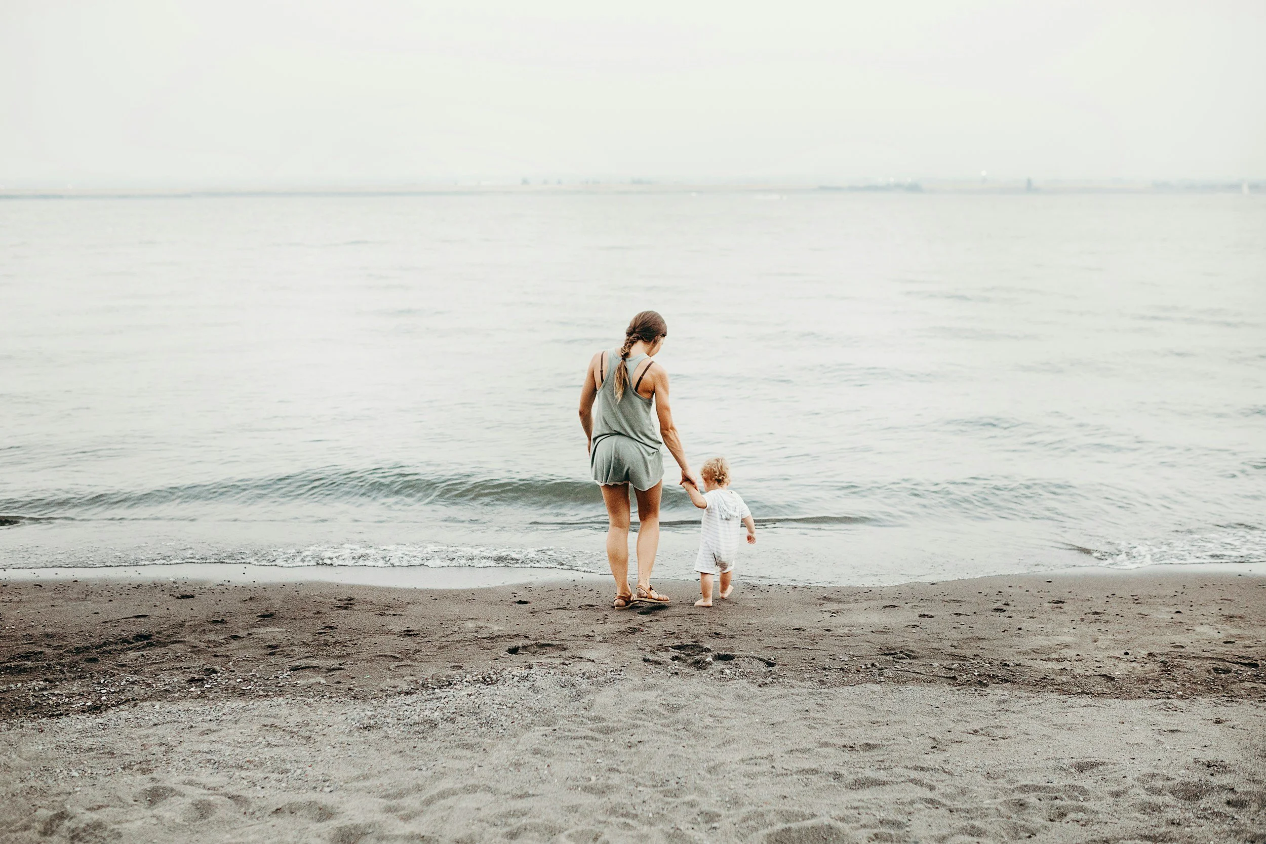 A woman and a young child walking hand-in-hand on the beach toward the water, with a grey sky and distant horizon.