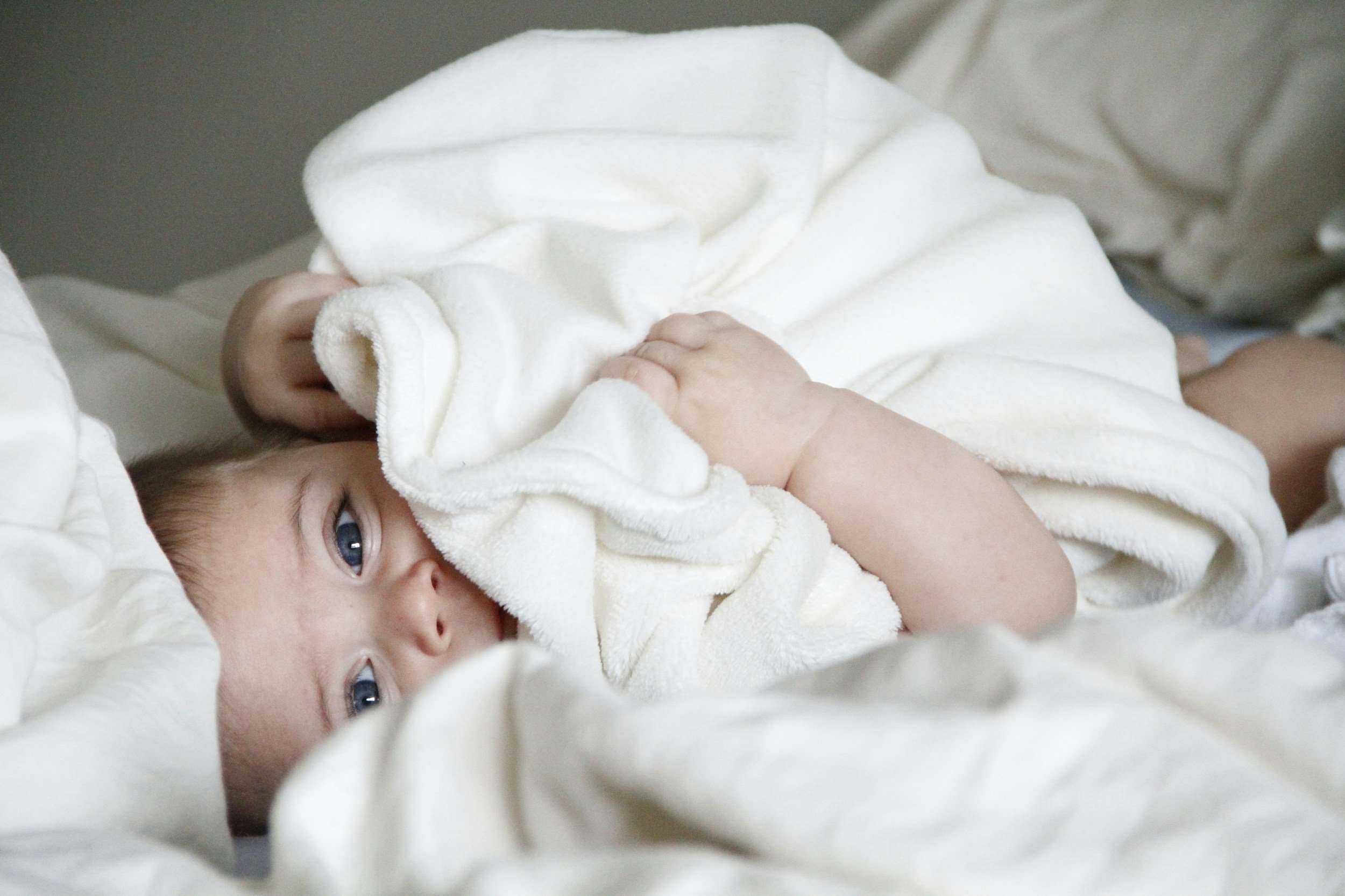 A baby lying on white blankets, partially covered, looking at the camera.
