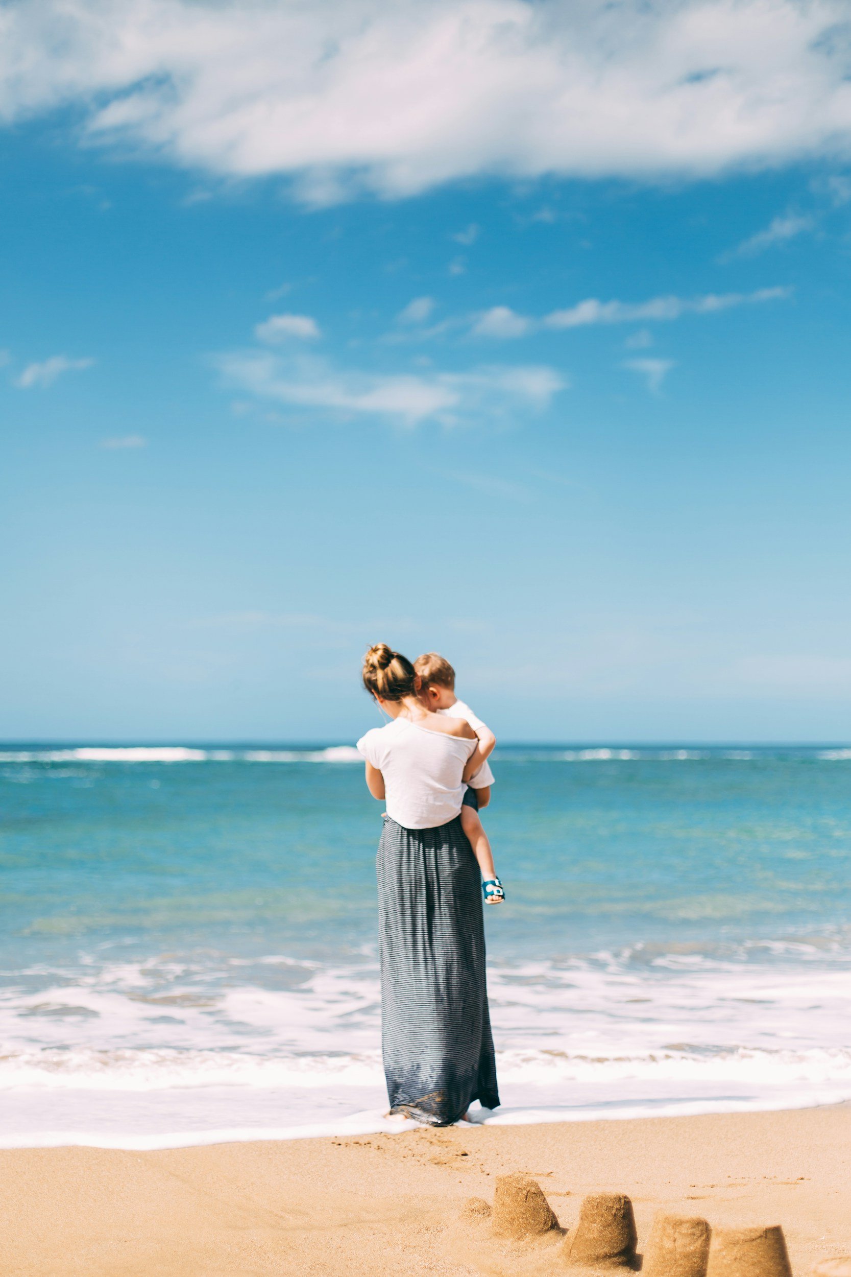 A woman holding a child on a sandy beach with the ocean in the background and a mostly clear blue sky.