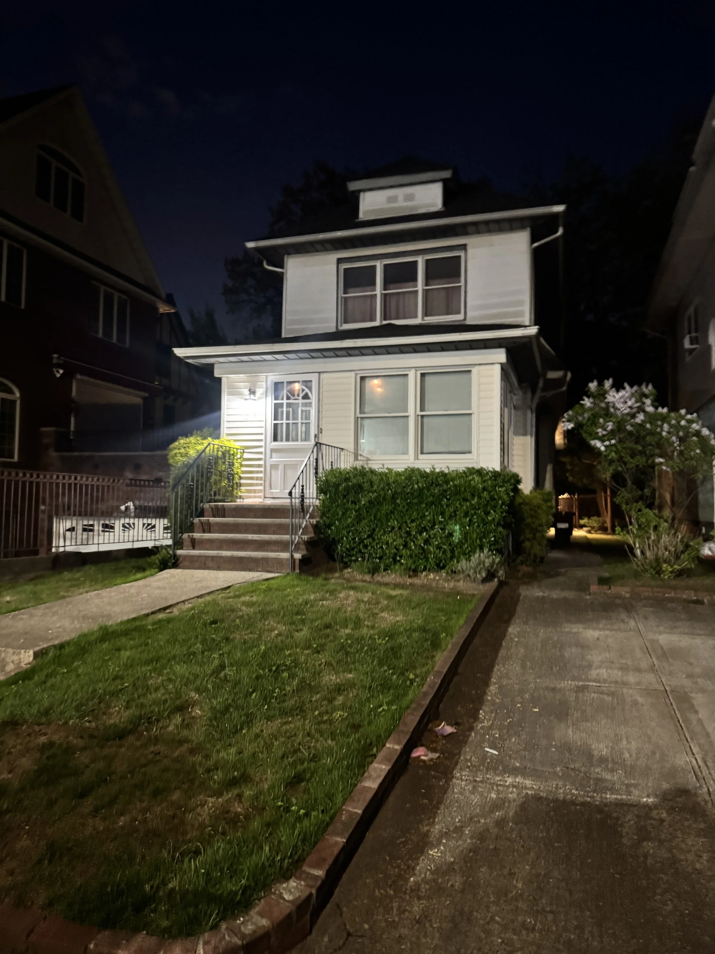 A two-story house at night with a front porch, stairs, and well-lit windows, surrounded by greenery and neighboring houses.