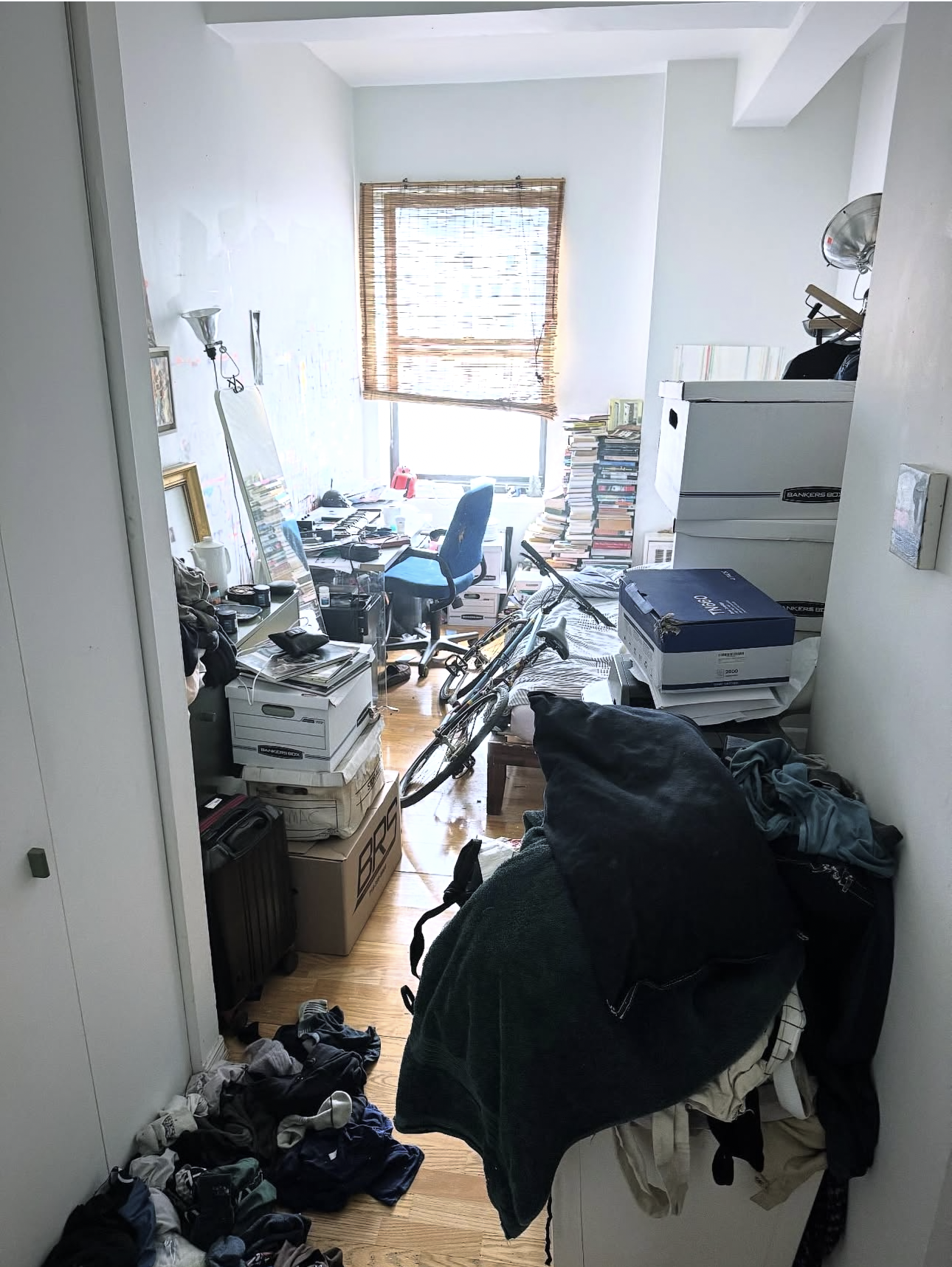 Messy bedroom with clothes on the floor, bike, and cluttered desk near window with blinds