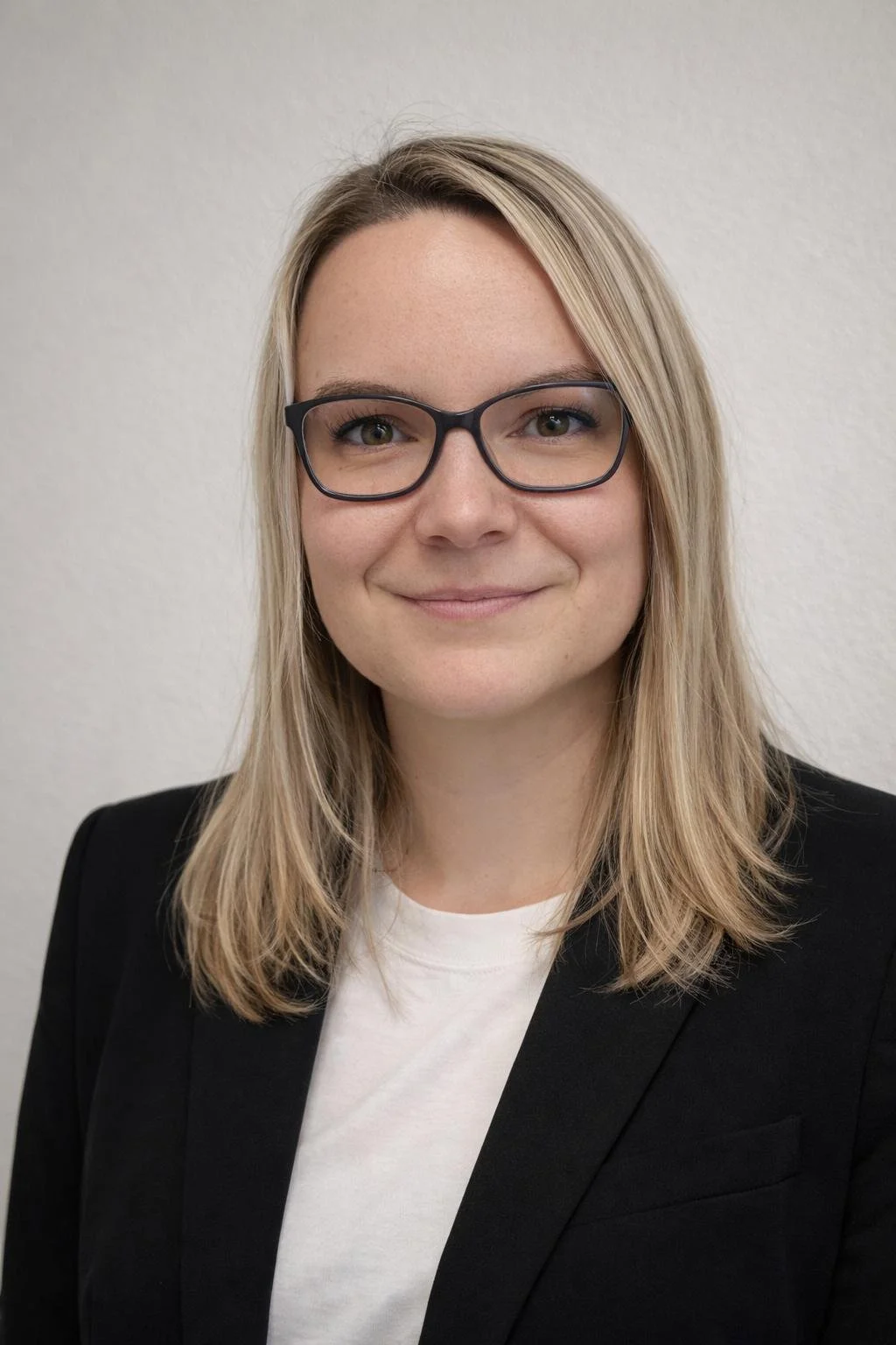Porträt einer lächelnden Frau mit Brille, blondem Haar und Business-Kleidung vor weißer Wand.
