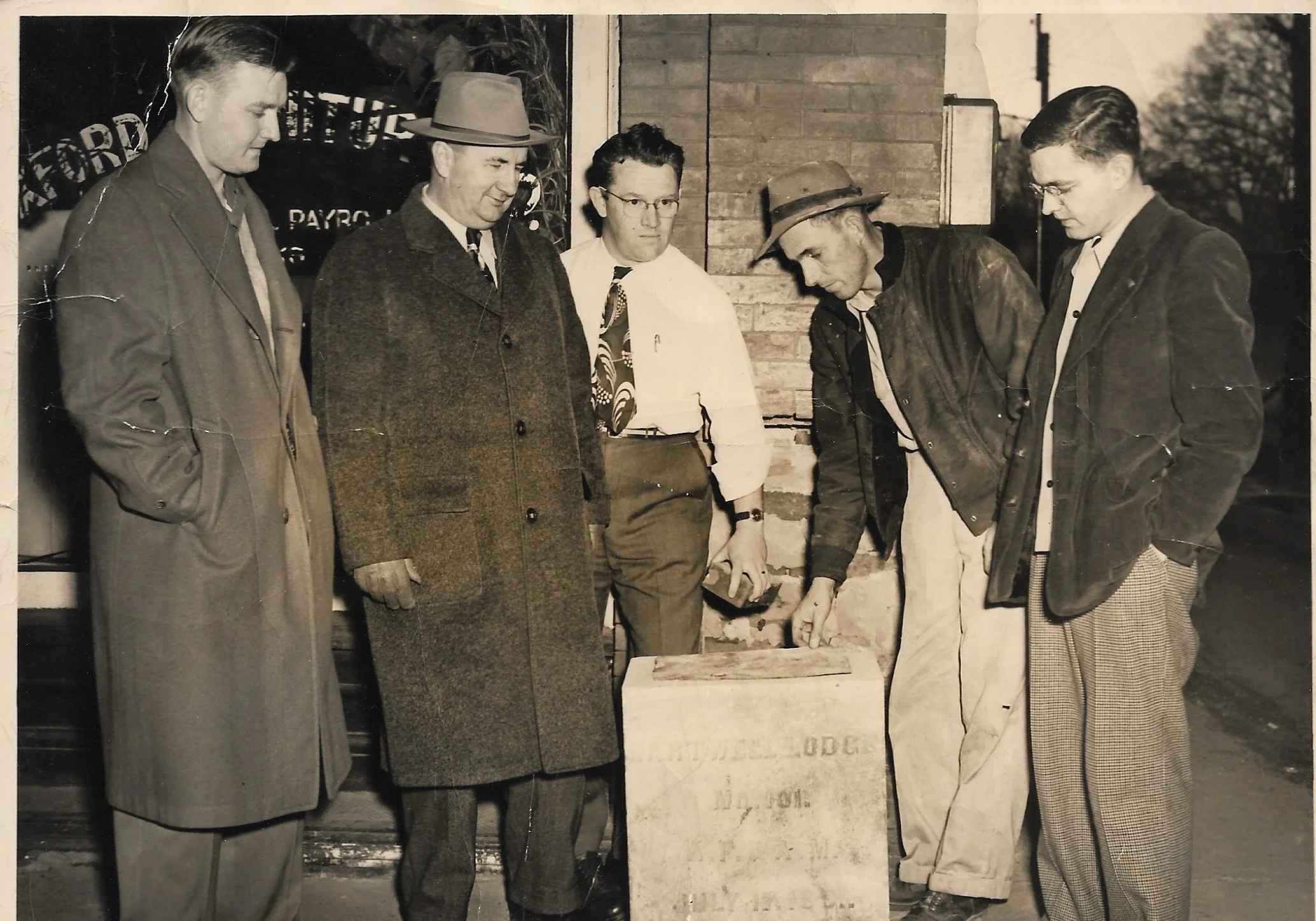 Six men in vintage clothing standing outdoors, gathered around a small stone monument, with some looking at it closely. The scene appears to be from mid-20th century, possibly during the 1940s or 1950s.