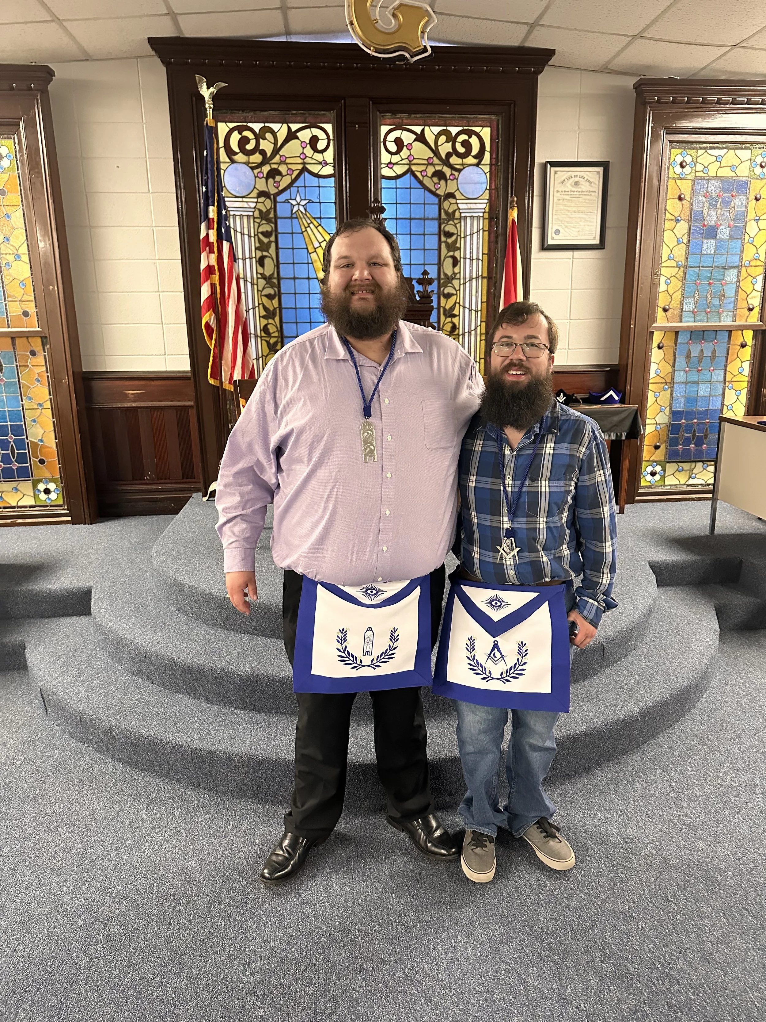 Two men with beards and glasses standing together in a Masonic lodge. They are wearing Masonic aprons and medallions, with stained glass windows and flags in the background.