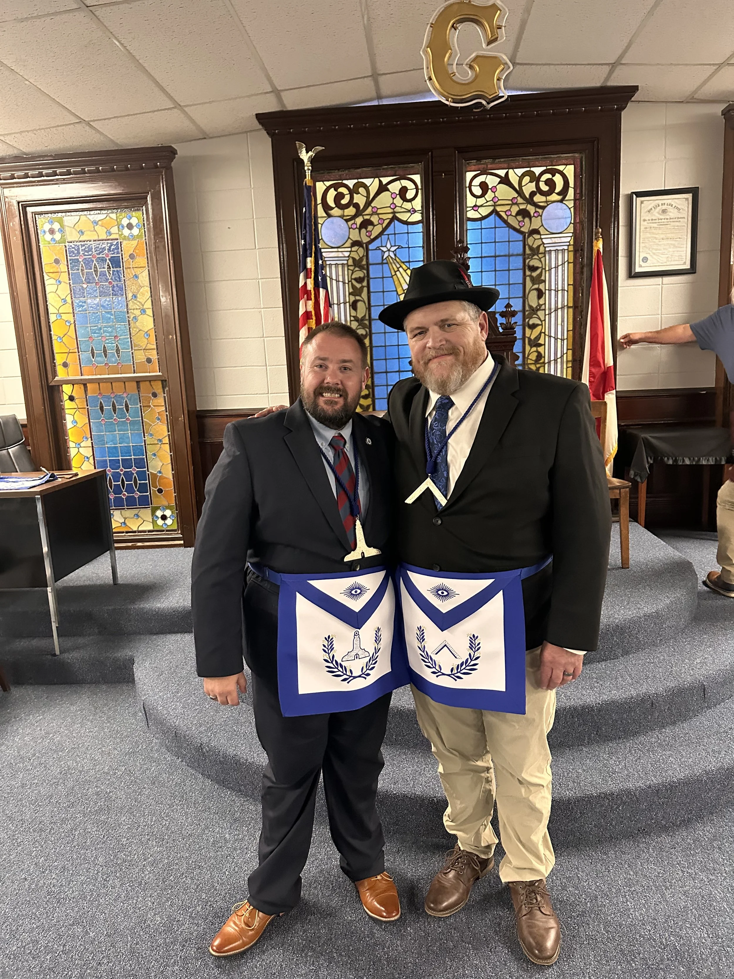 Two men in Masonic regalia standing side by side in a formal room with stained glass windows, American flag, and a decorative G symbol overhead. They are smiling and dressed in suits, with aprons and collars signifying Masonic fraternal roles.