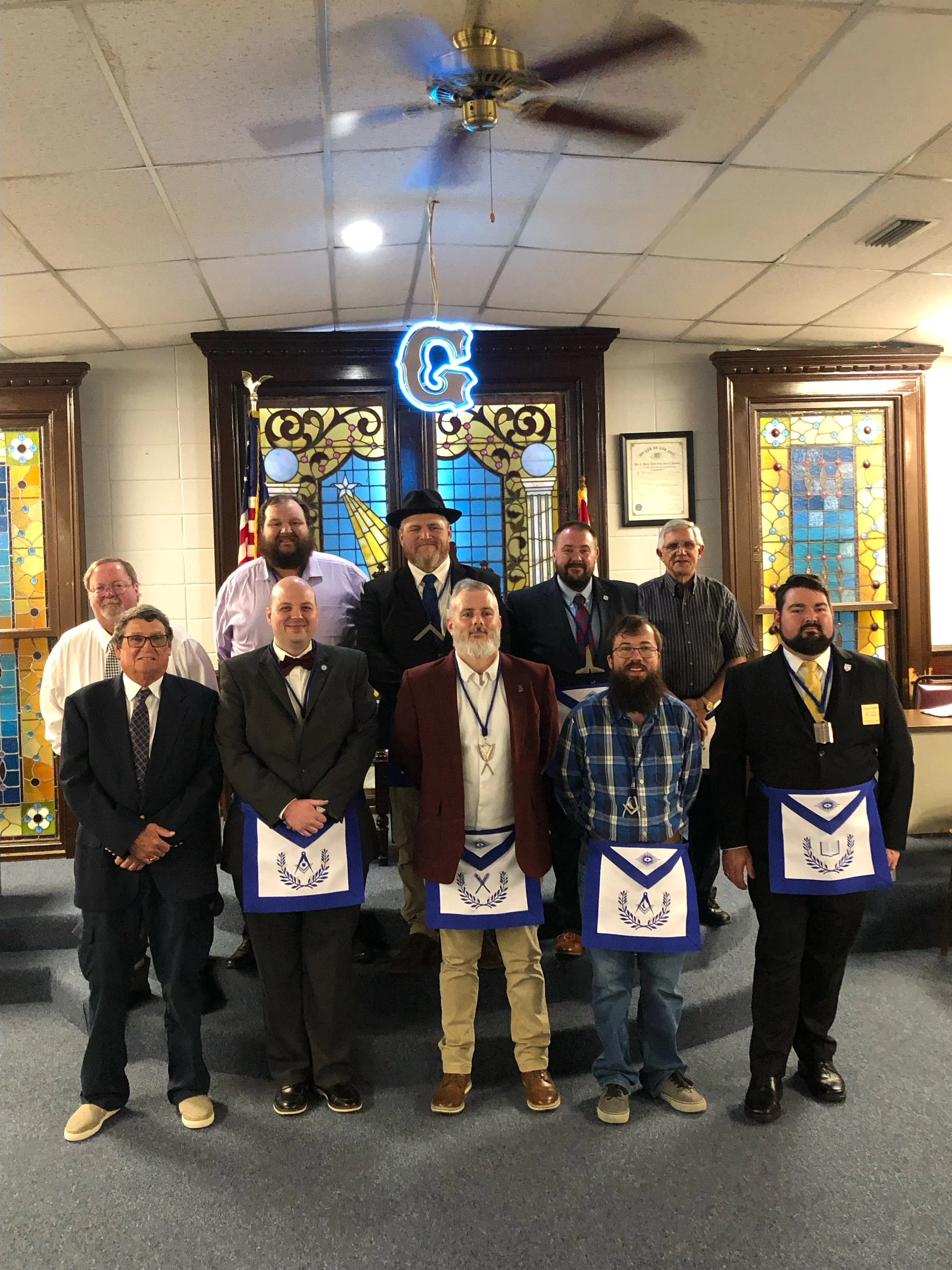 Group of ten men standing in a Masonic lodge, some wearing aprons with Masonic symbols, in front of stained glass windows and a blue letter 'G' hanging from the ceiling.