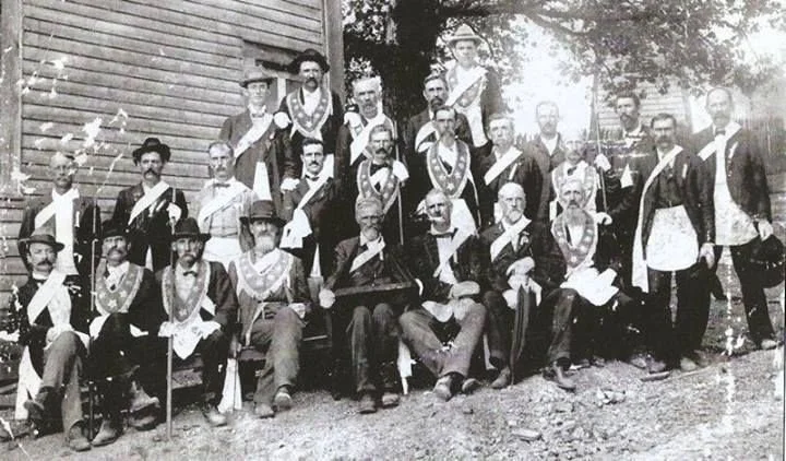 A black and white photo of a group of men in historical military or ceremonial uniforms. They are posed outdoors in front of a wooden building, with some seated and some standing, and appear to be part of a formal or ceremonial gathering.
