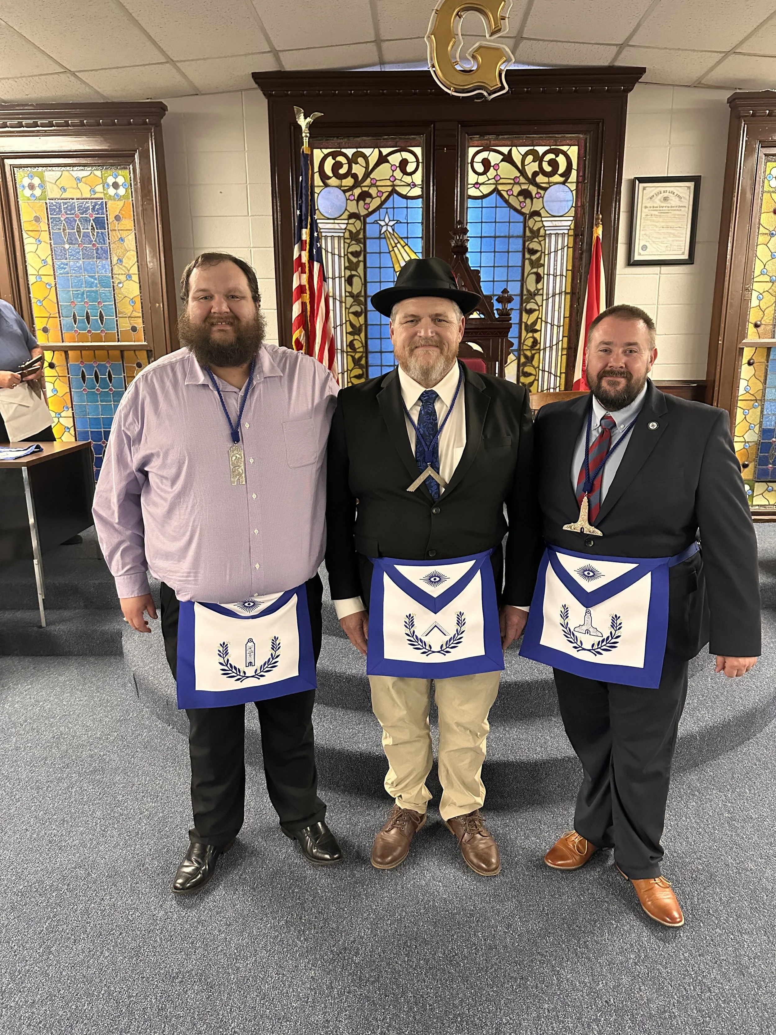 Three men standing in a room with stained glass windows, wearing aprons with Masonic symbols, and the man in the middle is wearing a black hat and a black suit.