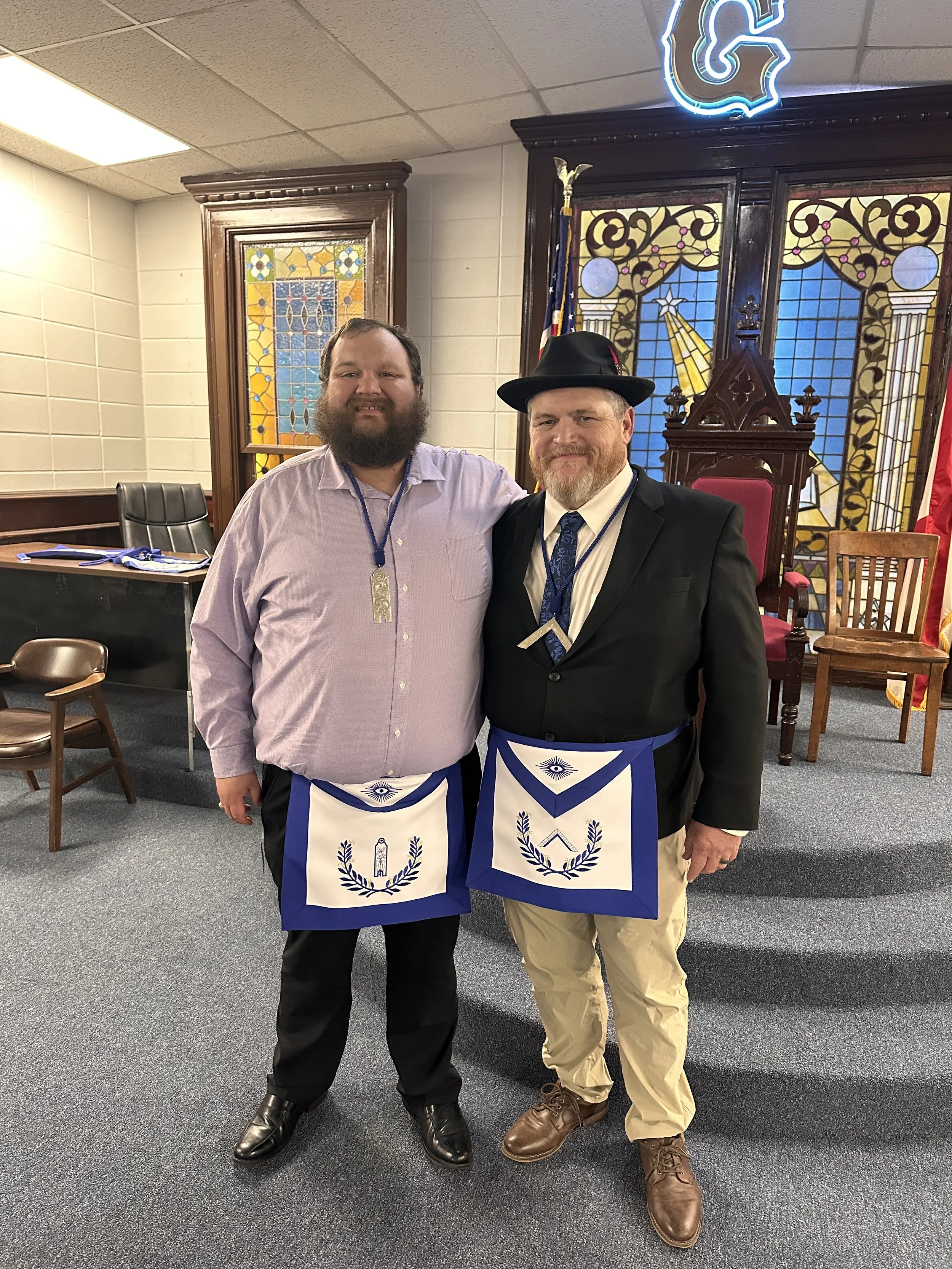 Two men standing side by side in a room with stained glass windows and an Masonic setting, both wearing aprons with Masonic symbols and clothing fitting a Masonic ceremony or gathering.