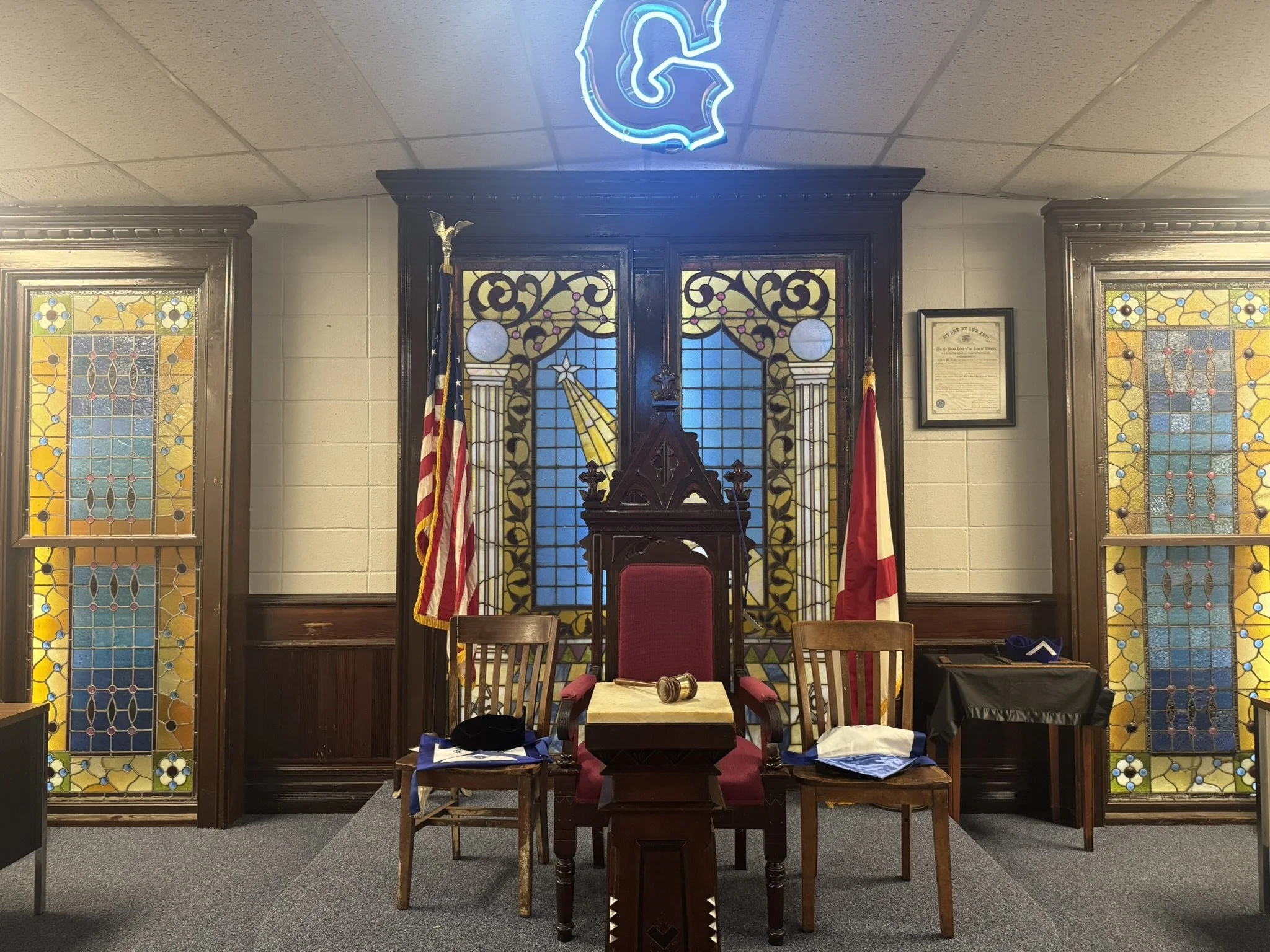 A Jewish synagogue's interior with a central seating area featuring a large red velvet chair with a gavel on the armrest, flanked by two wooden chairs with cloth on the seats, stained glass windows with blue, yellow, and brown patterns, Jewish and Am