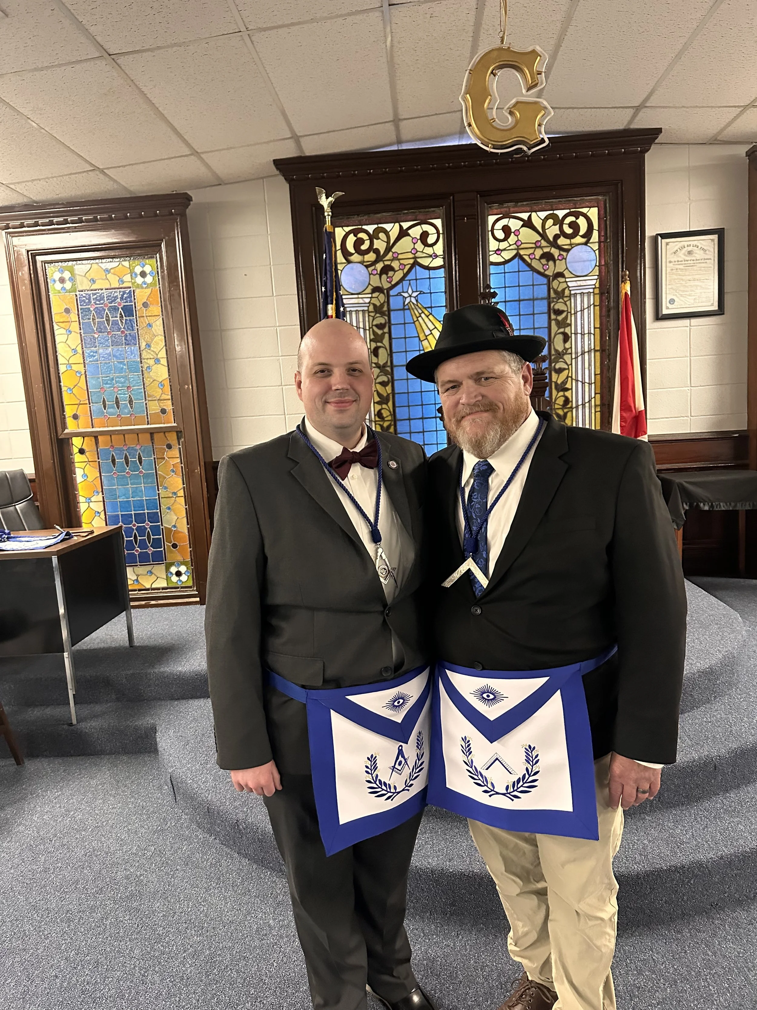 Two men in formal attire wearing Masonic aprons and neck ribbons standing inside a room with stained glass windows, a flag, and a large golden letter 'C' hanging from the ceiling.