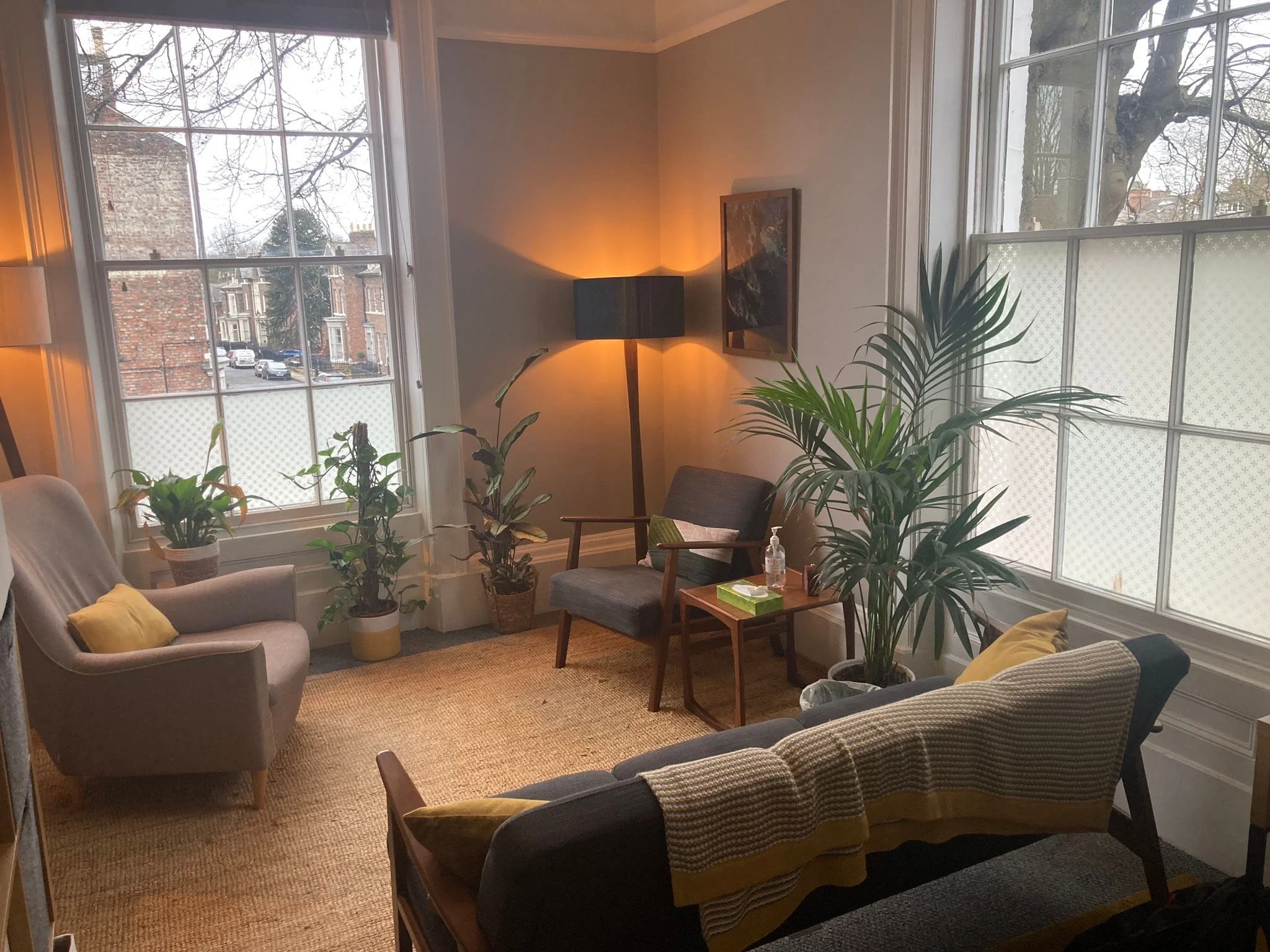 Living room with large windows, beige walls, a gray armchair, a patterned beige sofa with yellow pillows, a small wooden side table with hand sanitizer and tissue box, a floor lamp, a framed painting, and several potted plants.