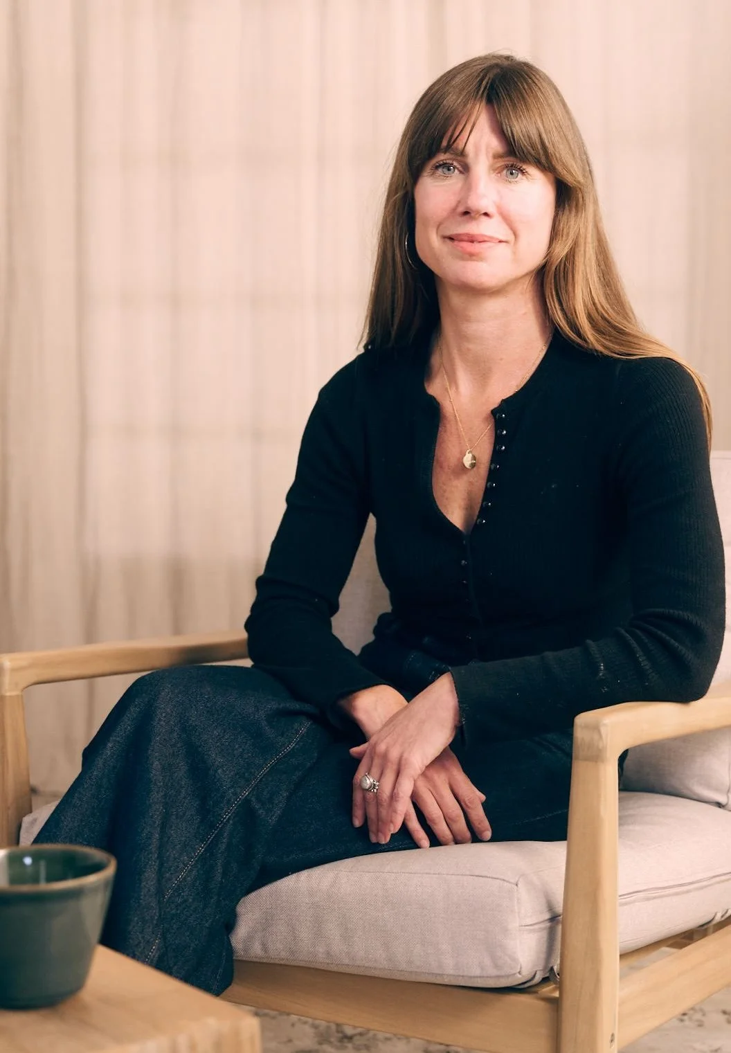 A woman with long brown hair wearing a black top, sitting on a beige cushioned chair, looking at the camera with a neutral expression, in a room with beige curtains.