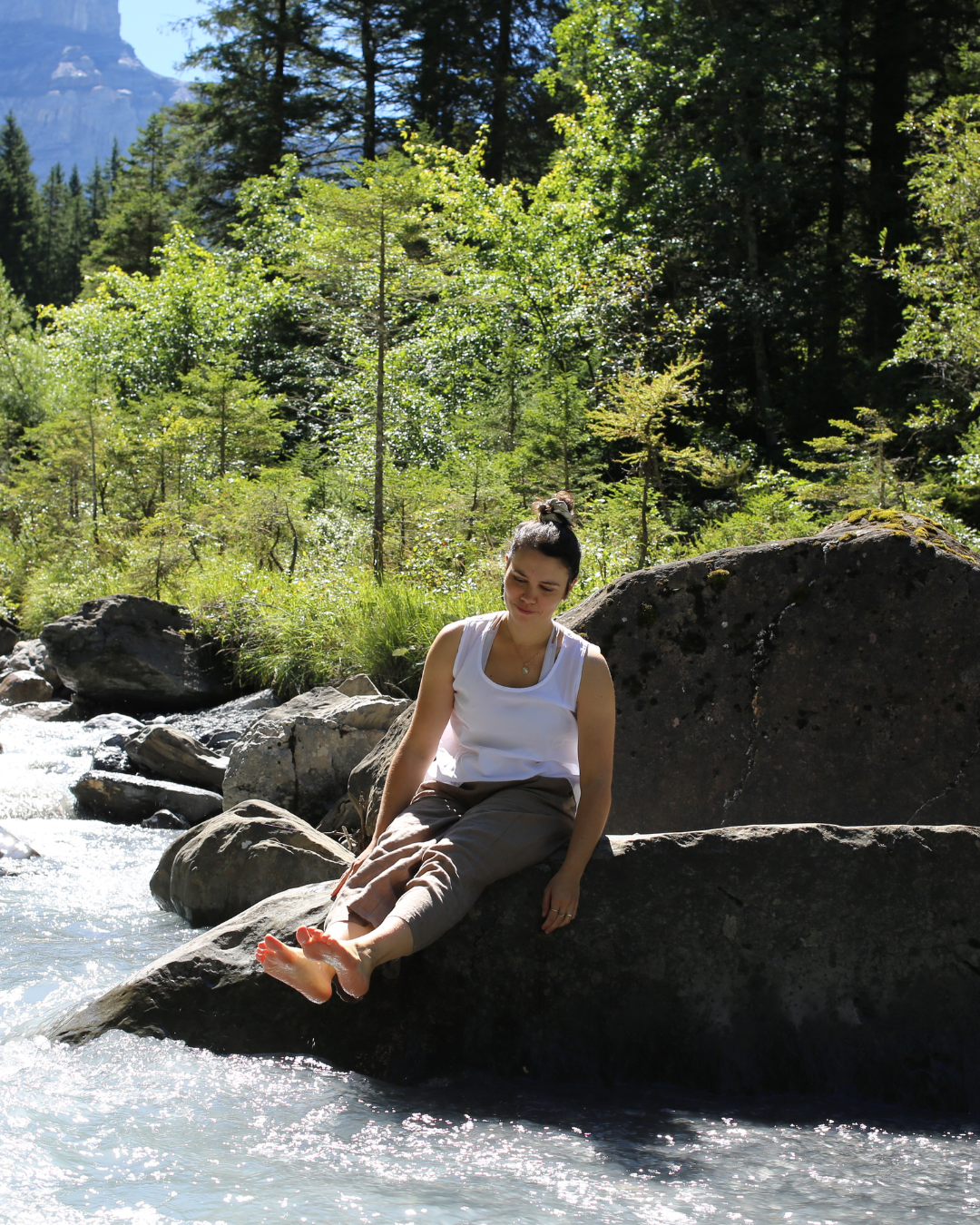 Une femme assise sur un rocher au bord d'une rivière en forêt, portant un haut blanc et un pantalon beige, avec des cheveux attachés en haut, entre des arbres verts sous un ciel bleu.
