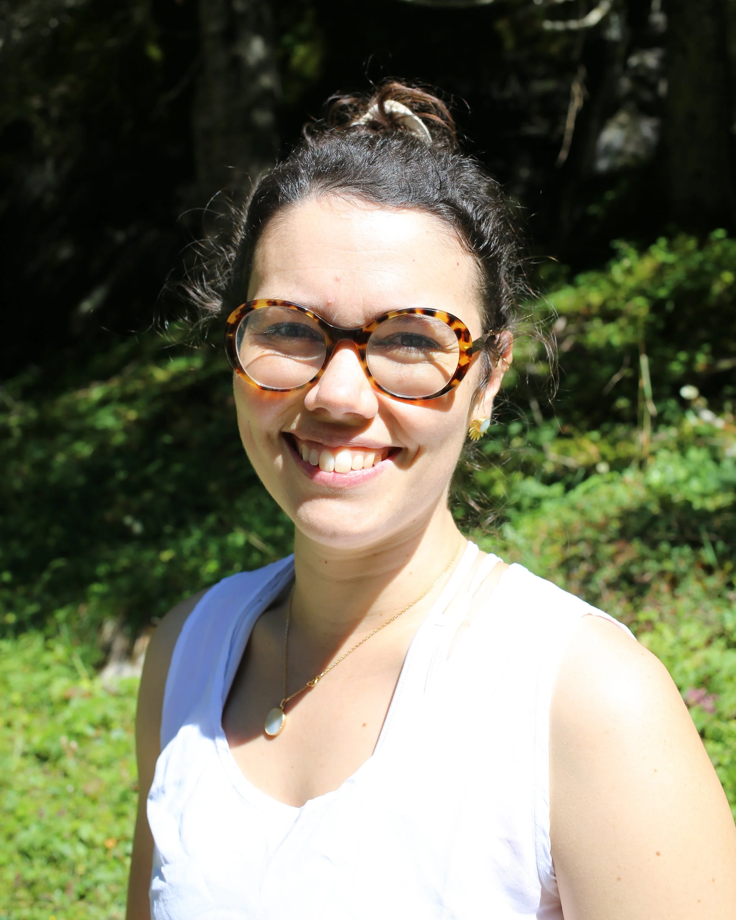 Jeune femme souriante portant des lunettes écaille et un collier, posant à l'extérieur avec un arrière-plan de verdure sous une lumière solaire vive.