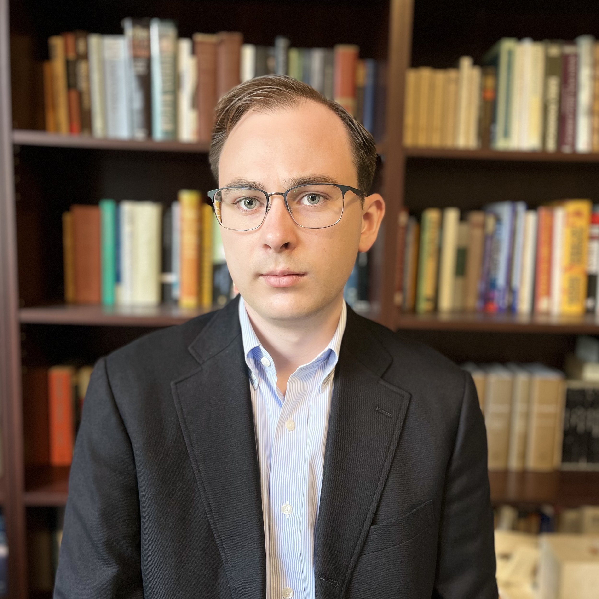 A man with glasses, wearing a suit and light blue shirt, standing in front of a bookshelf filled with books.