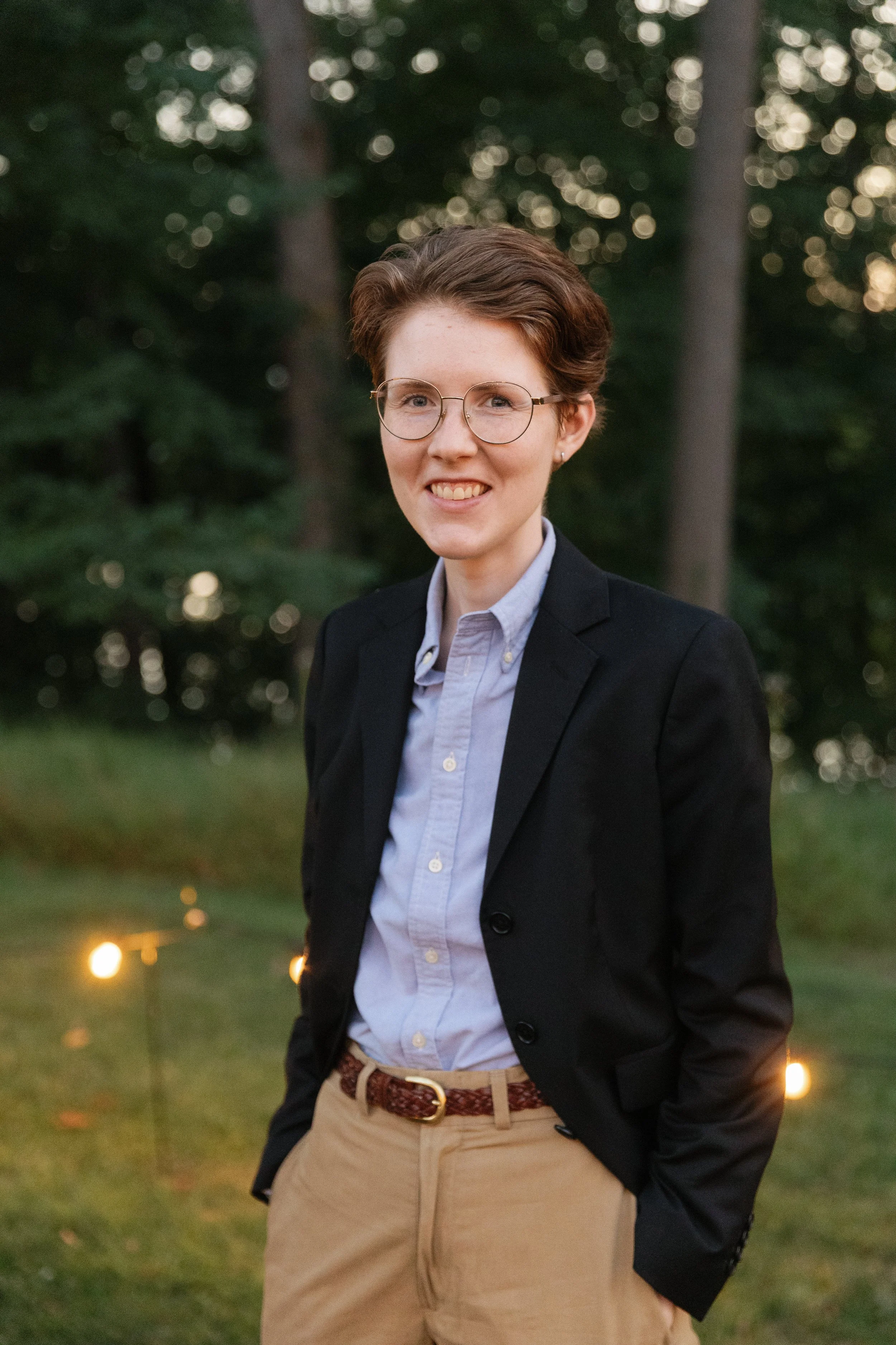 A young person with short brown hair, glasses, and a bright smile, dressed in a dark blazer, light blue shirt, and beige pants, standing outdoors with trees and string lights in the background during sunset.