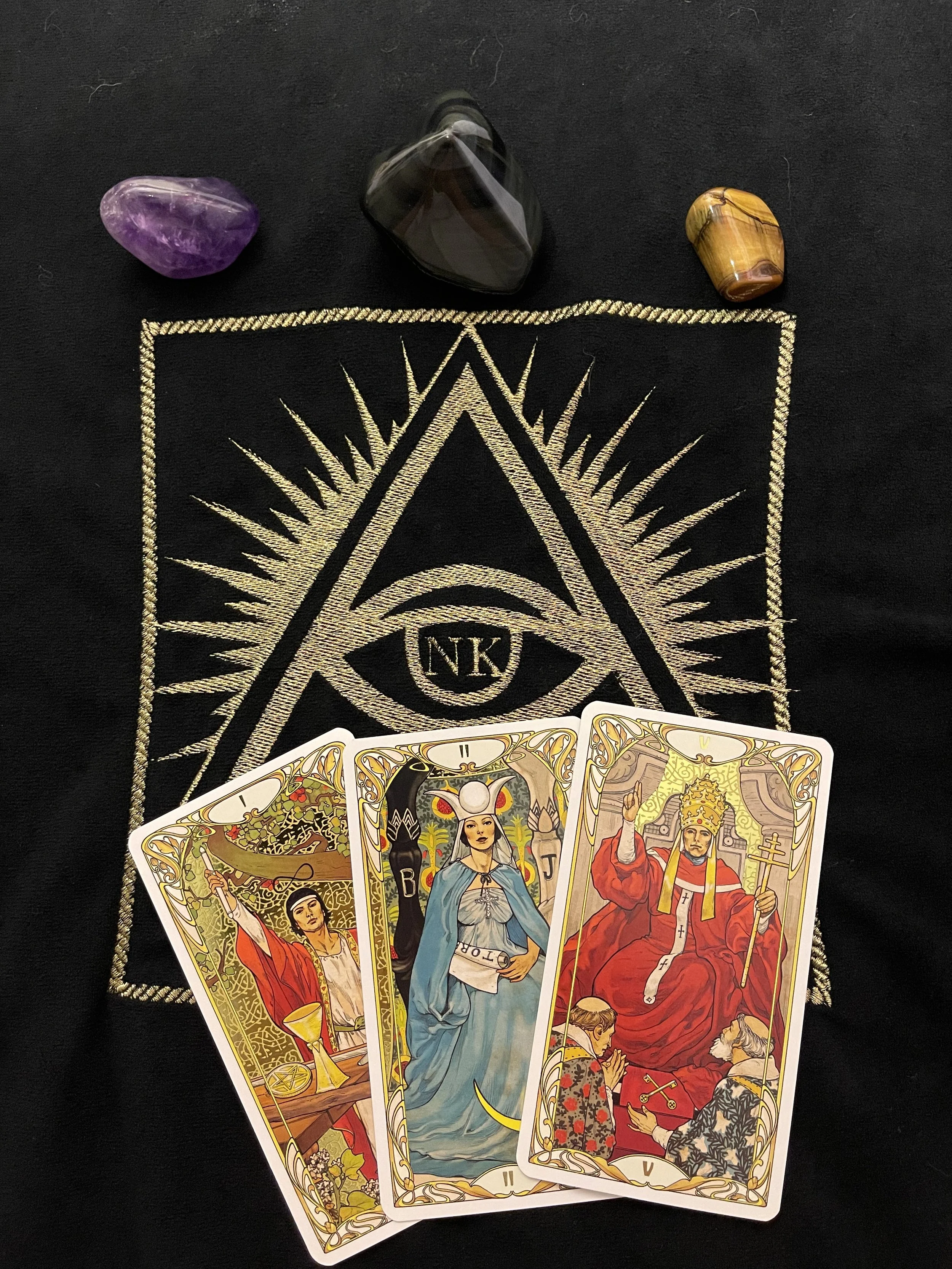 Trois cartes de tarot disposées sur un tissu noir avec un symbole d'œil dans un triangle et un rayon de lumière doré, accompagnées de trois pierres précieuses (améthyste, onyx,Œil de tigre).