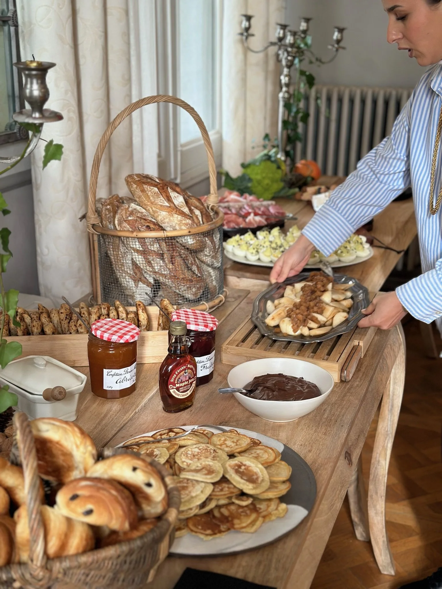 Table de petit-déjeuner avec divers pains, confitures, gaufres, fruits, sauces, œufs, charcuterie et décorations dans une ambiance chaleureuse.