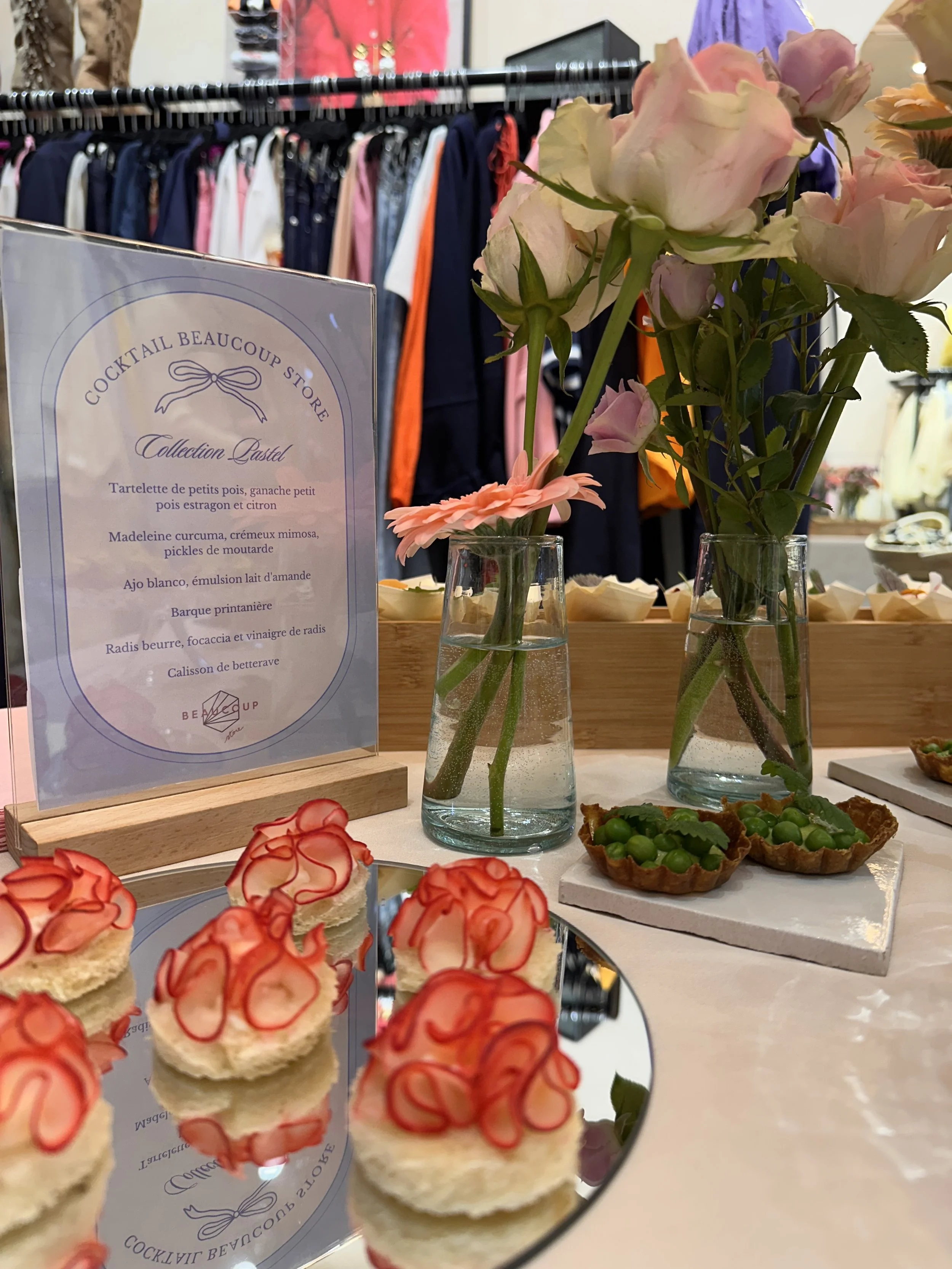 Vitrine avec roses roses dans deux vases, une assiette avec petits cupcakes décorés de coulis de fraise, et deux tartes avec des pois verts, sur une table dans une boutique.