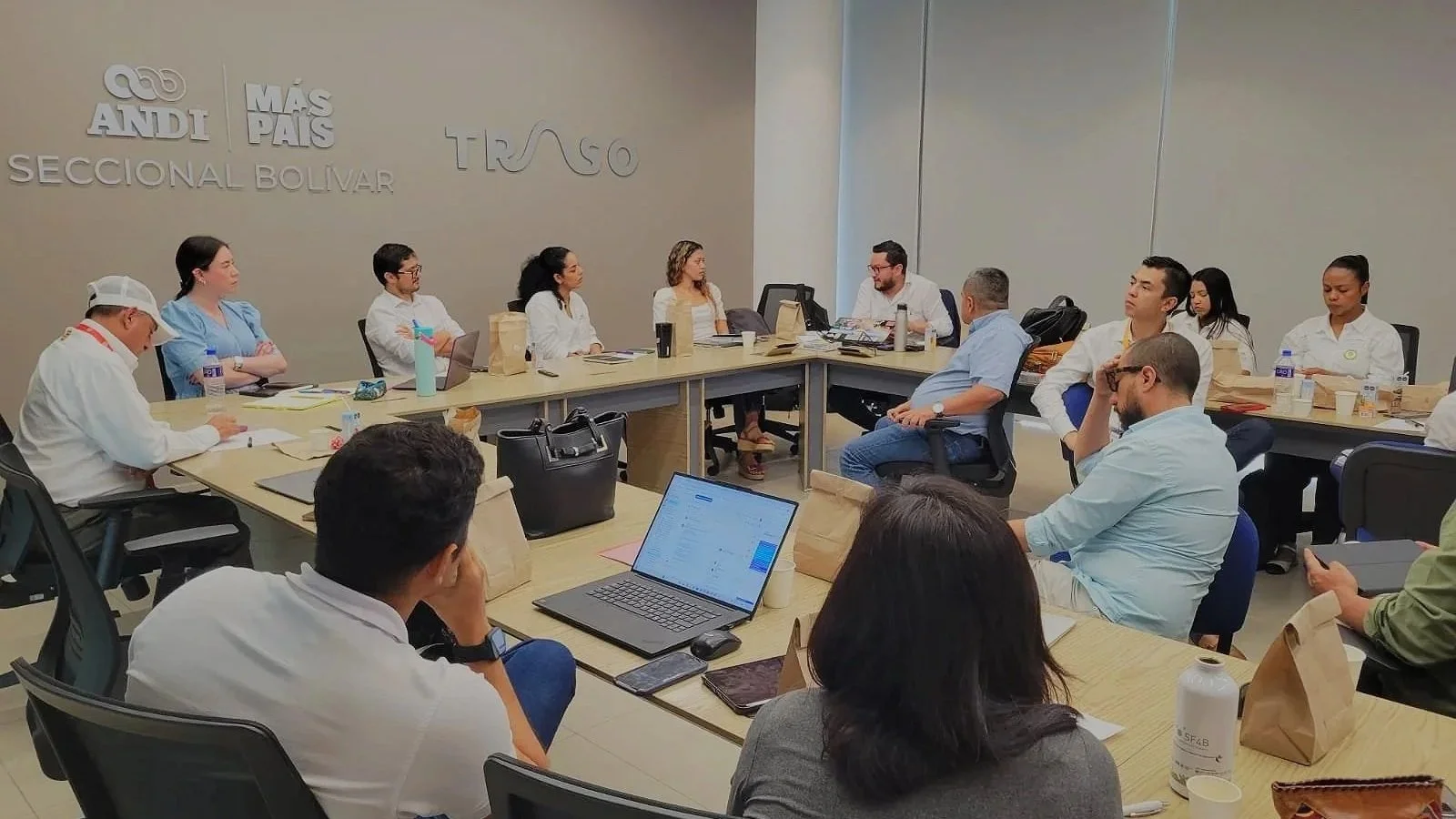 Grupo de personas en una reunión en una sala de conferencia, sentados alrededor de una mesa larga con computadoras, papeles y bebidas, en un ambiente profesional.