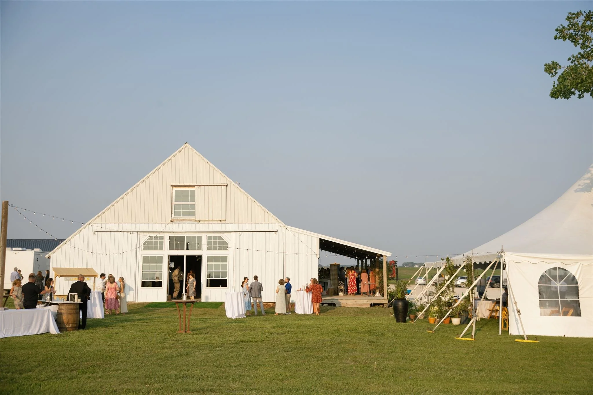 white barn, gabled roof, large windows, grassy area, people socializing, tables, large white tent