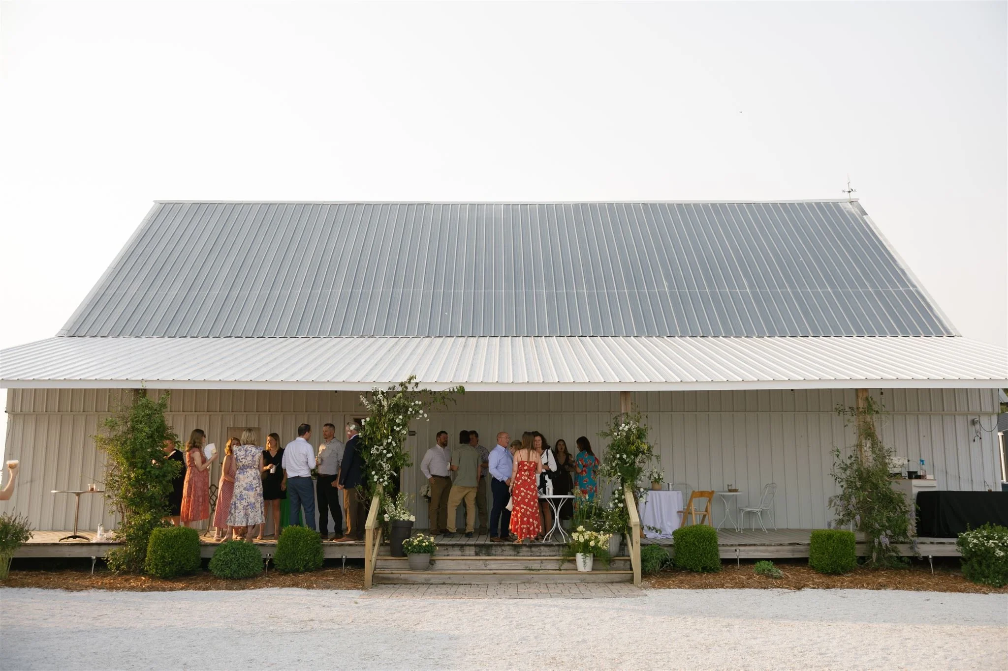 People gathered on porch outside white barn, decorated greenery, white flowers, wedding celebration.