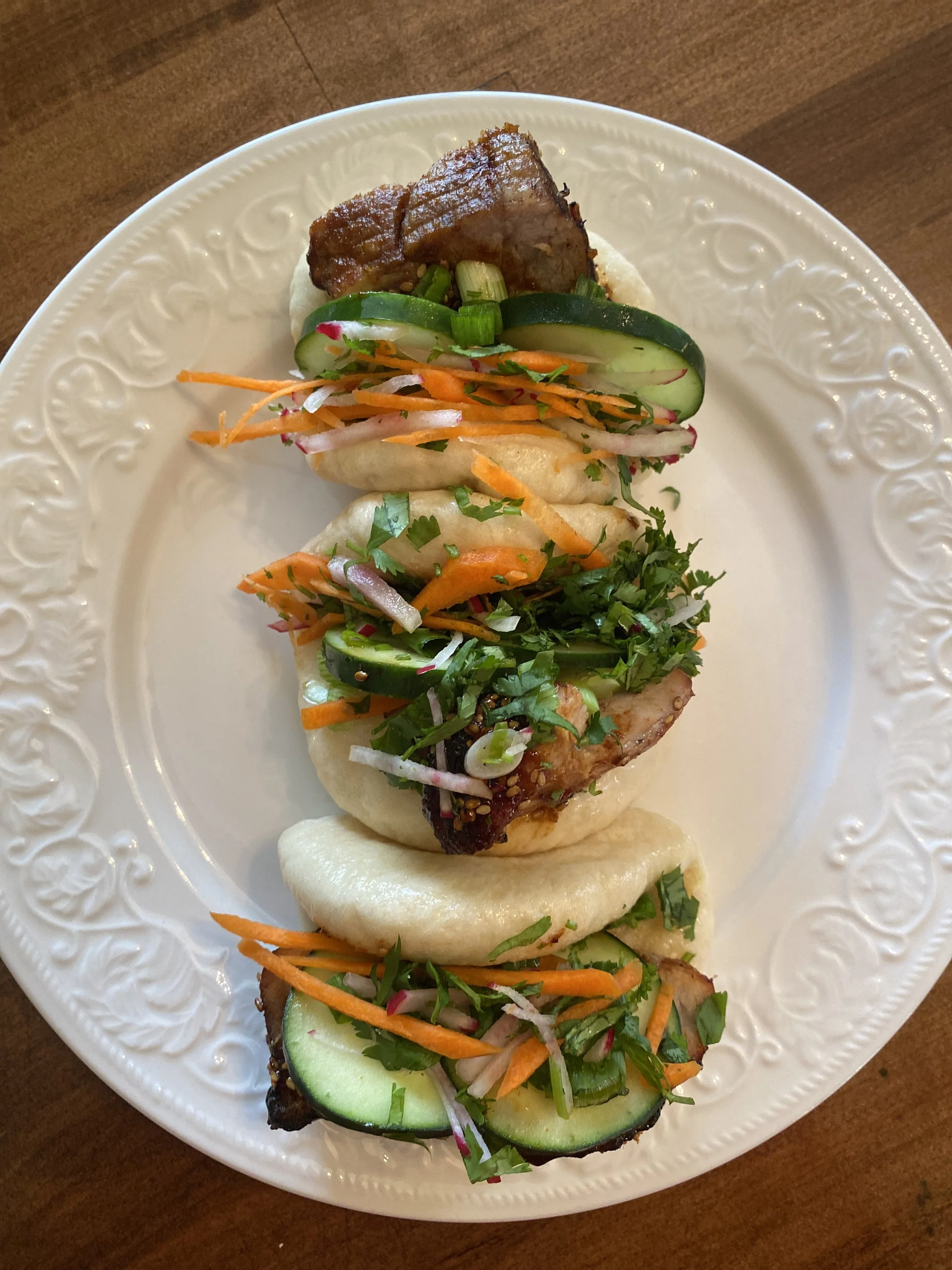 Plate of three pork bao buns topped with sliced cucumbers, shredded carrots, onions, fresh herbs, and vegetables.