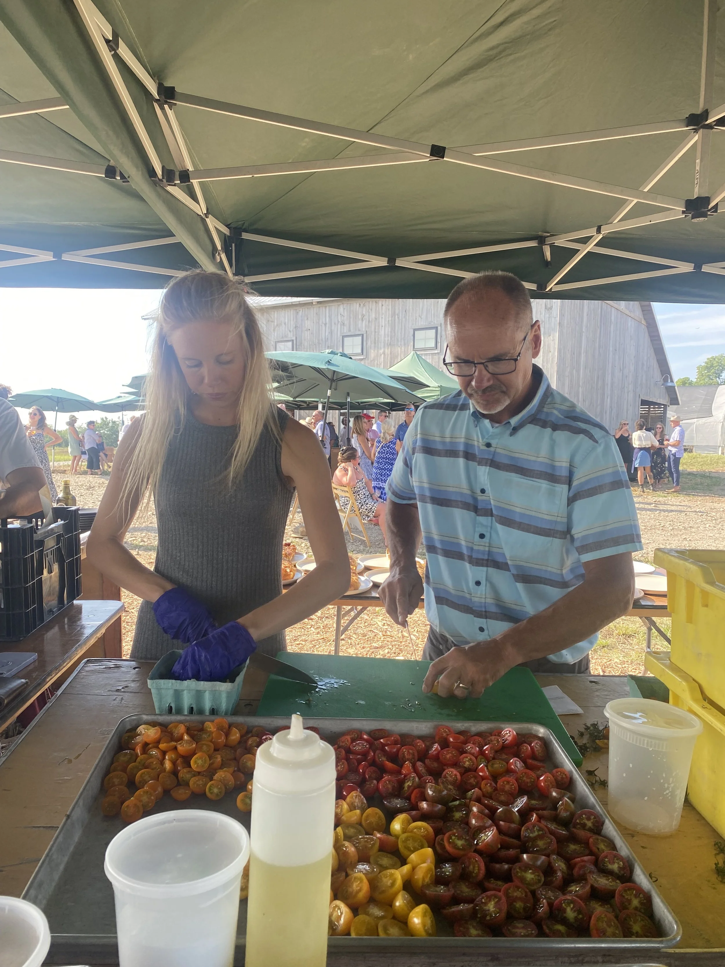 Two people cutting tomatoes, outdoor farm to table dinner, tray of mixed heirloom tomatoes.