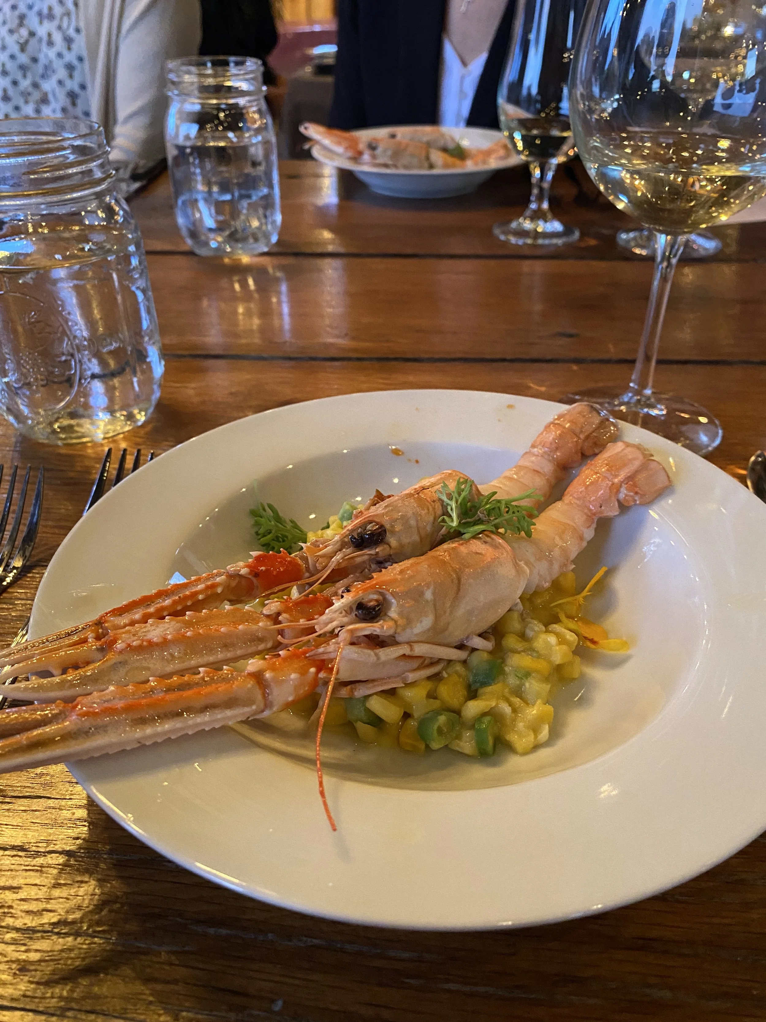 Cooked shrimp on a bed of mixed vegetables. Glasses of white wine, water, wooden table.