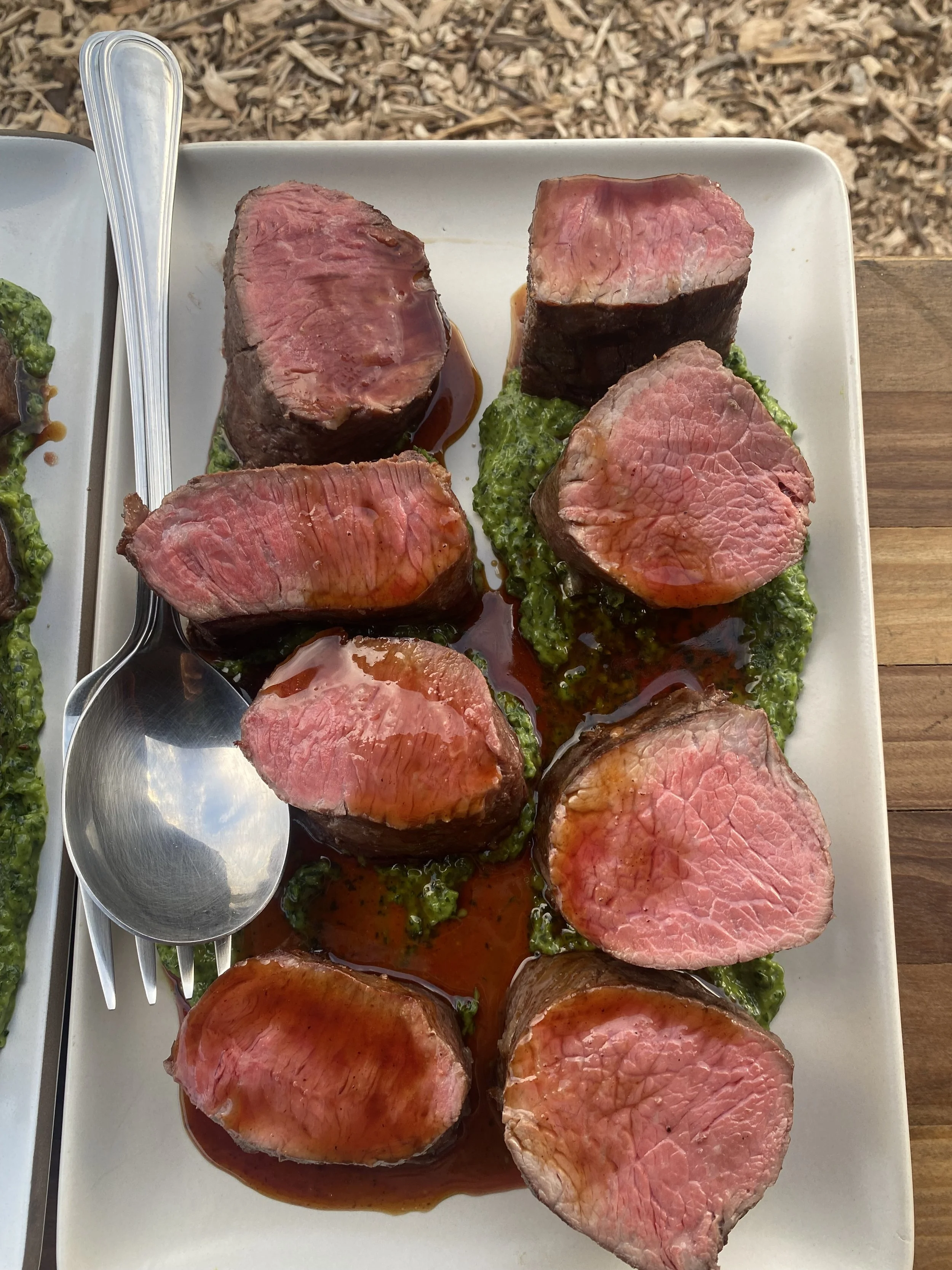 Sliced beef tenderloin topped with jus, white plate, green chimichurri.