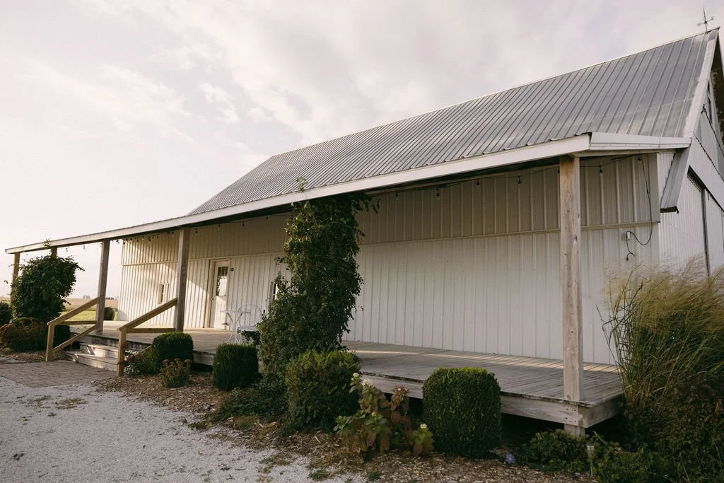 A rural landscape with a white barn, a large tree, a grassy field, and a dirt road during sunset with clouds in the sky.