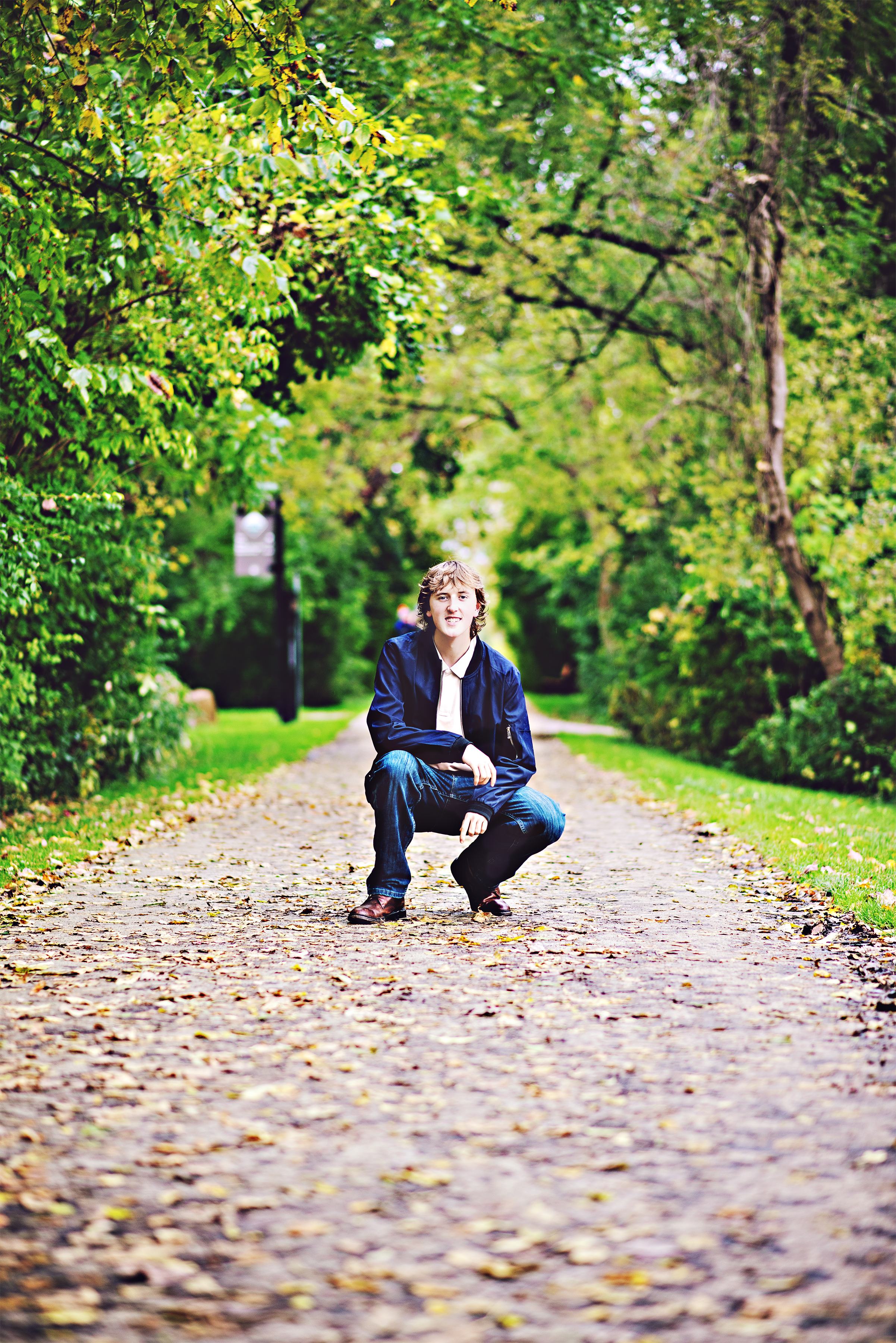 A young man crouching on a leaf-covered path in a park surrounded by trees with green foliage during daytime.