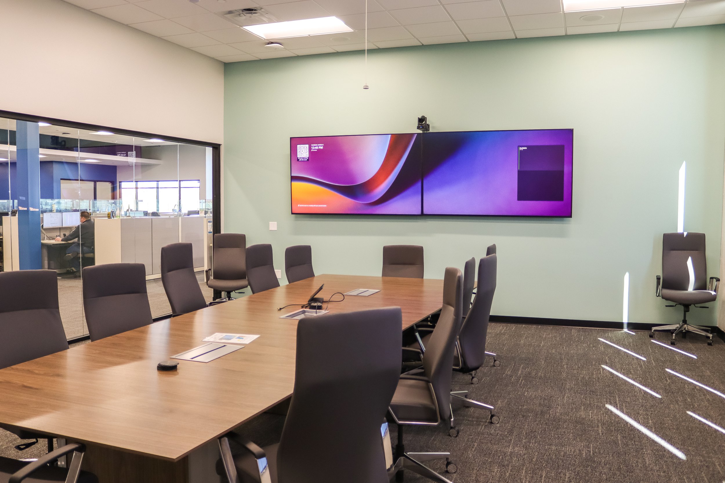 Modern conference room with a large wooden table surrounded by black office chairs, a large screen on the wall displaying a colorful background, and a glass wall revealing an office area with a person working at a desk.