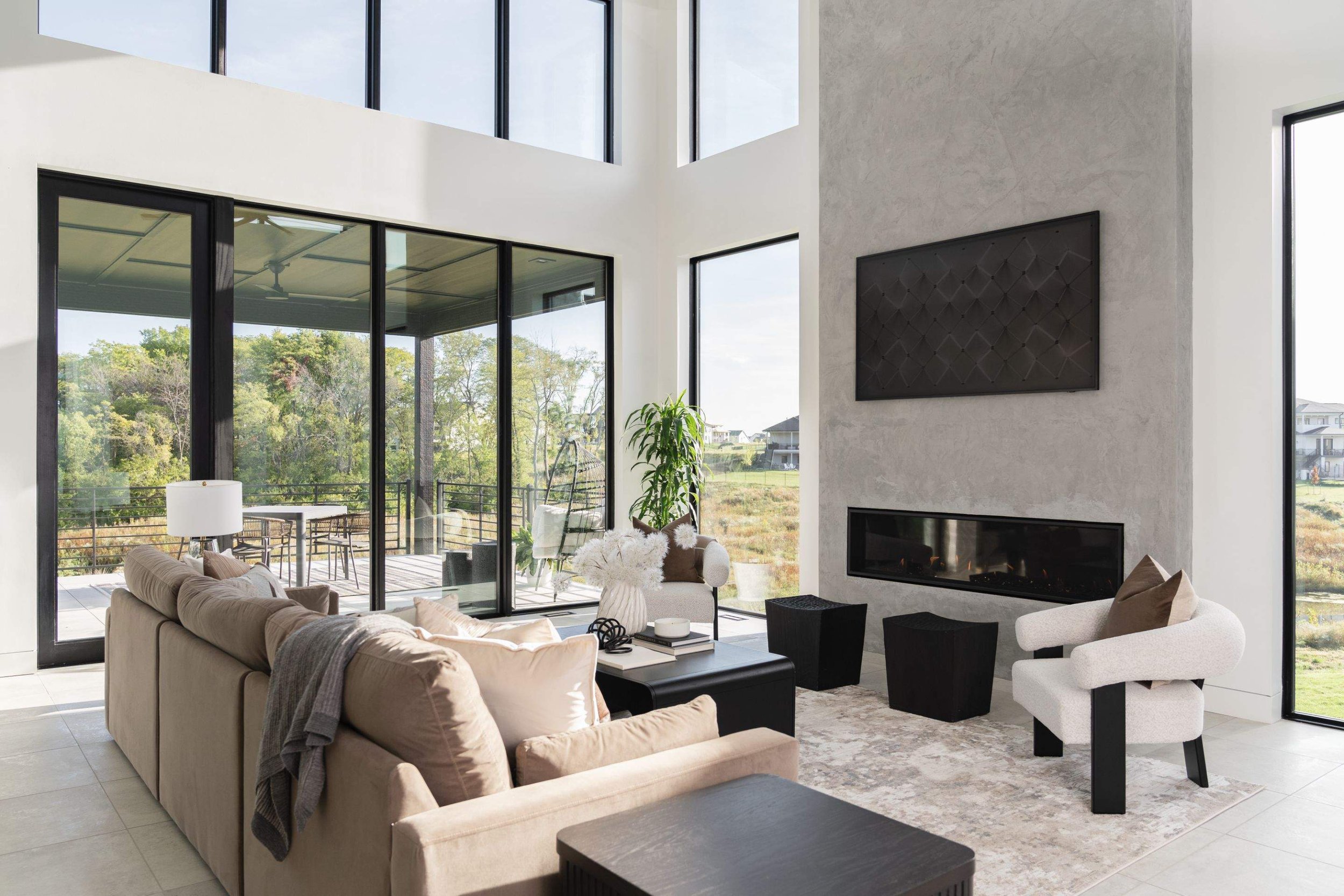 Modern living room with large floor-to-ceiling windows, a beige sofa, white armchair, black coffee table, and a fireplace on a gray wall.