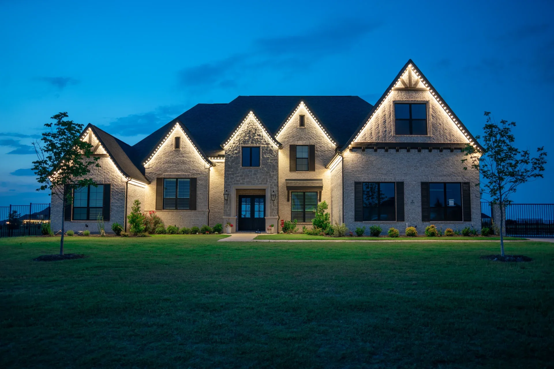 A large, illuminated house with multiple gabled roofs, brick and stone exterior, and decorated with string lights at dusk, surrounded by a green lawn and trees.